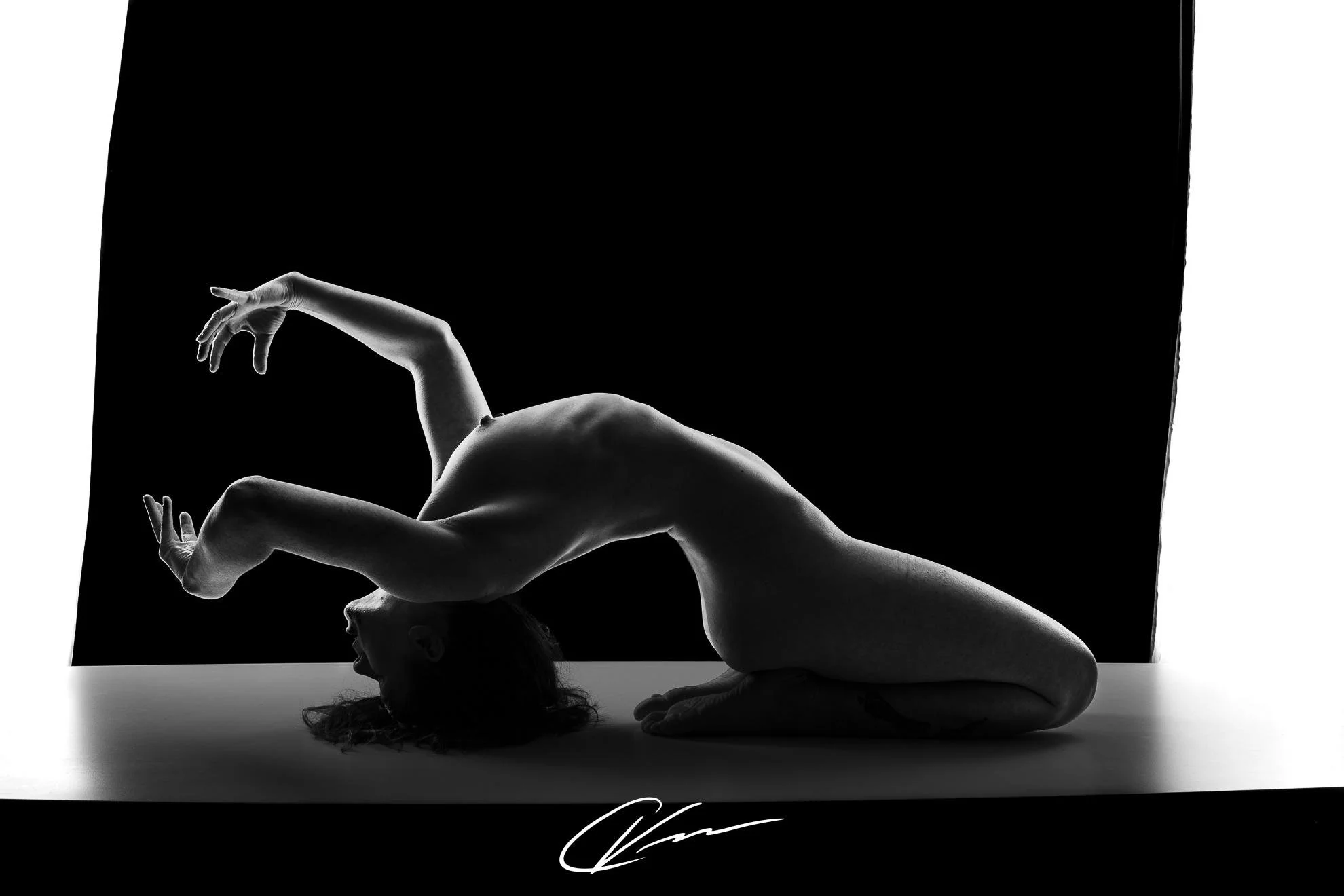 Black and white photo of a woman in a complex yoga pose, balancing on her knees and head with her arms extended, against a dark background.