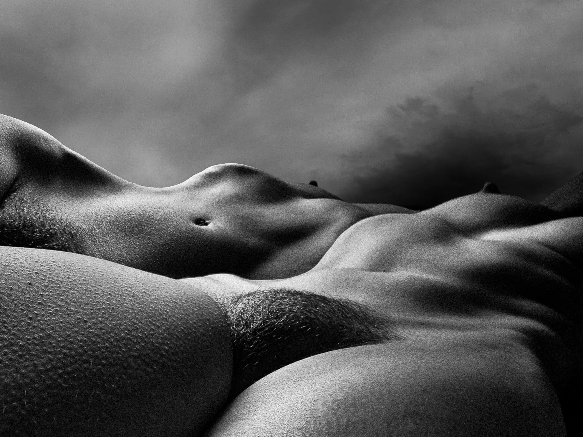 Black and white close-up photo of a nude torso, focusing on the chest, stomach, and side, with a cloudy sky background.