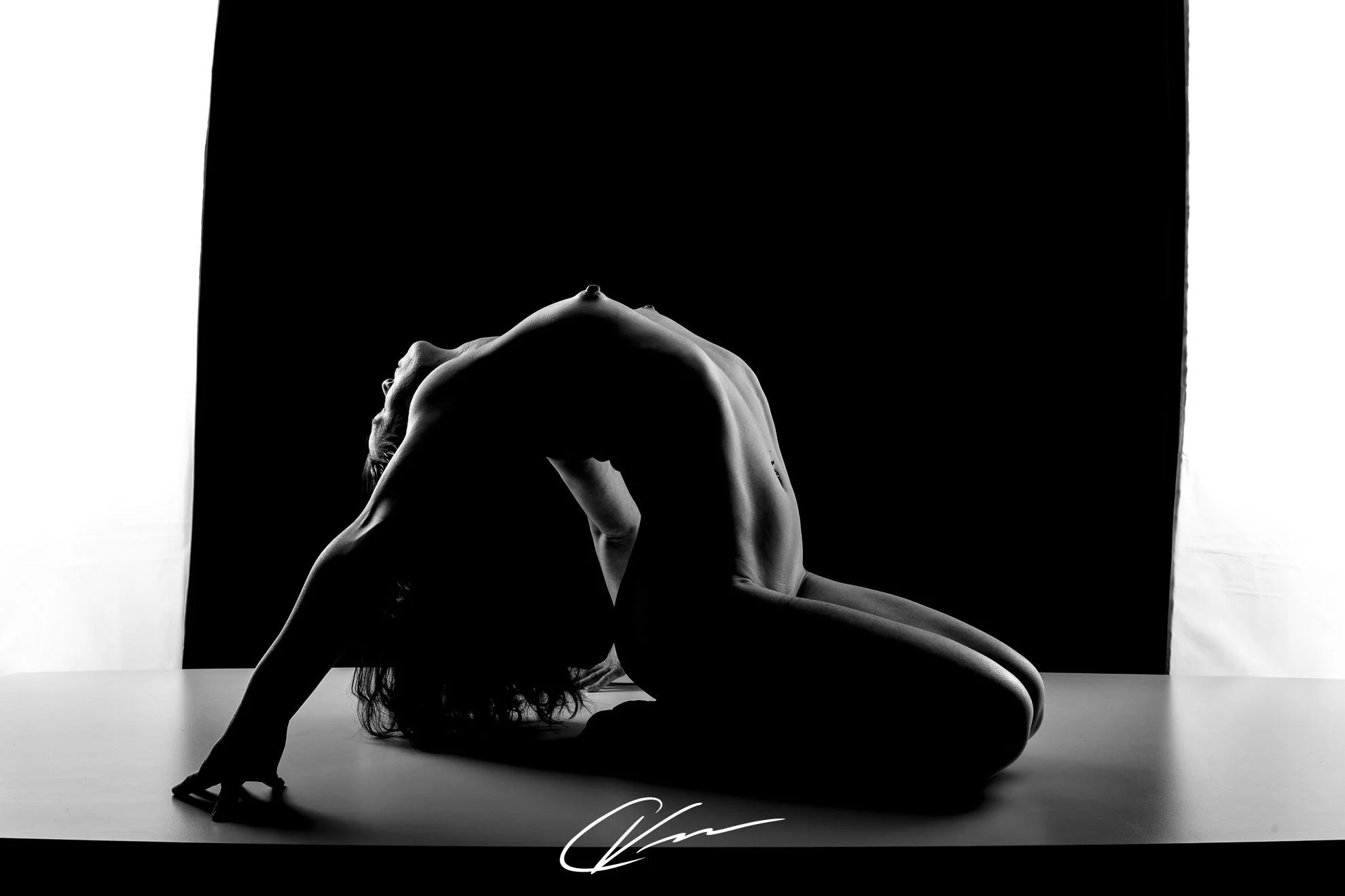 Silhouette of a nude woman in a kneeling yoga pose with her head down, back arched, and arms supporting her weight on the surface, against a dark background with white sides.