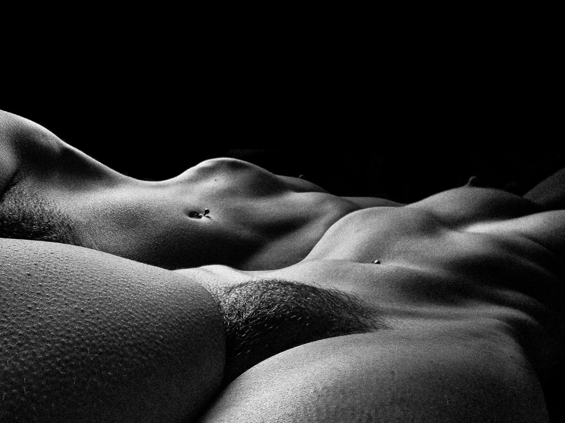 Black and white close-up of a topless woman lying down with her eyes closed and a small belly button piercing visible.