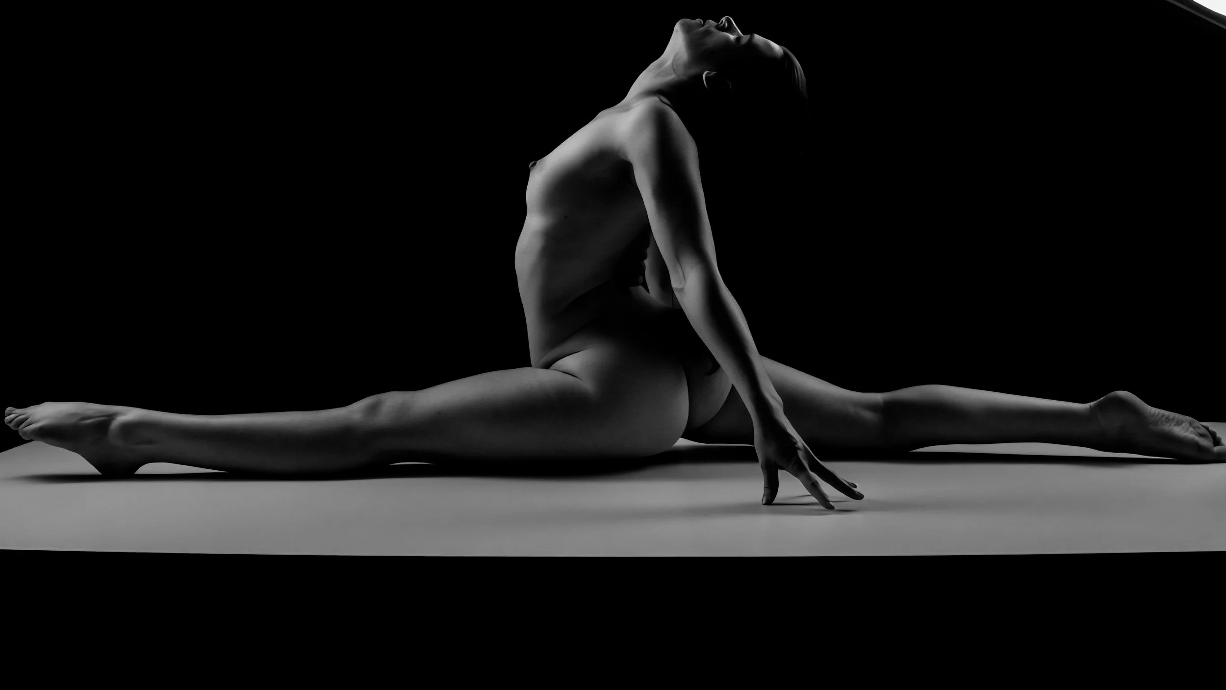 Black and white photo of a woman performing a split on the floor, with one hand placed on the ground and her head tilted back.