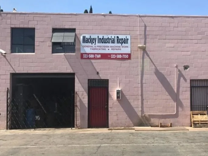 Pink industrial building with a sign that reads 'Mackey Industrial Repair' and contact information. The building has a small window, a red door, and some equipment and pallets outside.