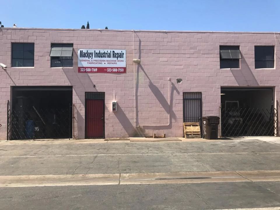 The building has a sign indicating it is Mackey Industrial Repair, specializing in general and precision machine work, fabricating, and repairs. The building is pink with small windows and a mix of garage and door entrances. There is a trash bin outside.