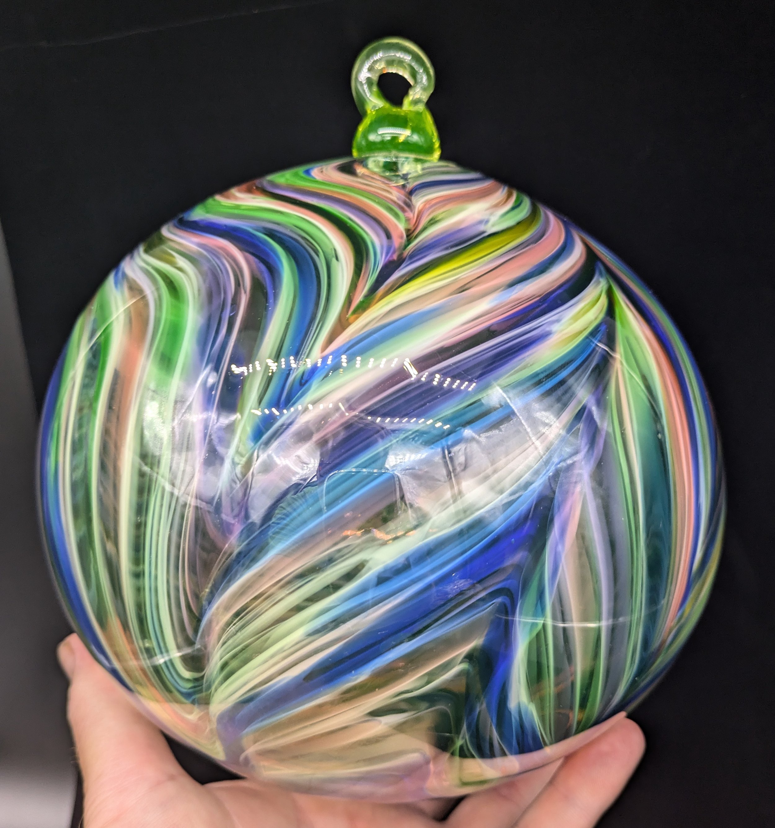 A hand holding a colorful uranium glass ornament with swirling patterns of green, pink, purple, and blue on a black background.