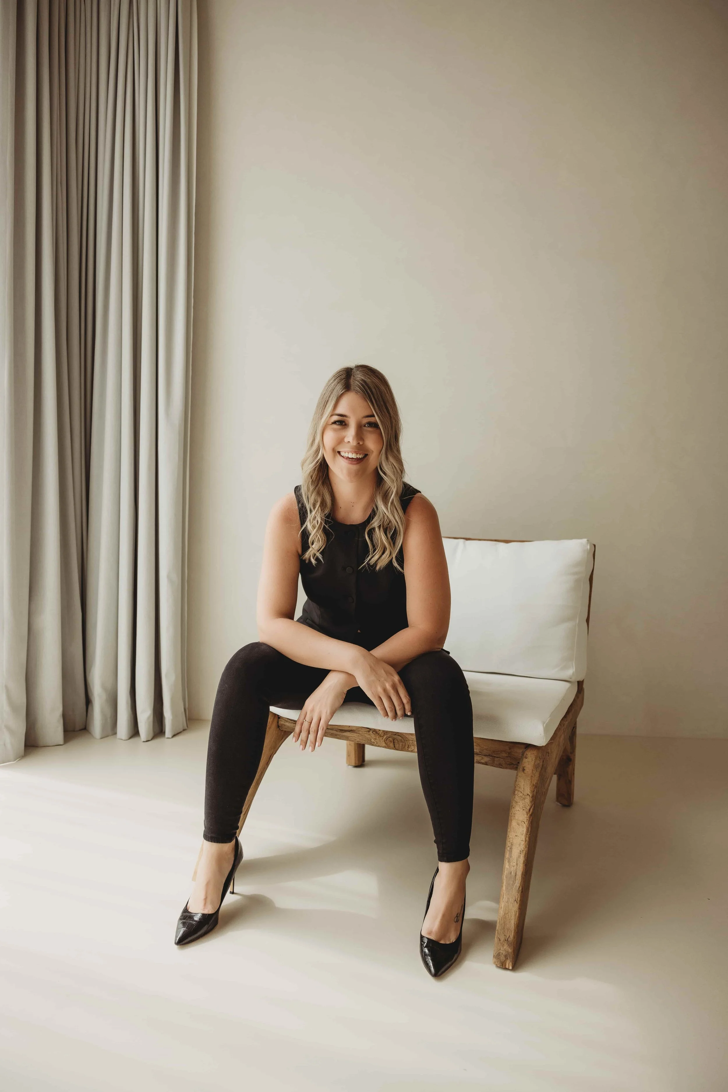 Kiara Jade Trotter in a black sleeveless top sitting on a wooden chair with her legs crossed, smiling in a minimalist room with white walls, beige curtains, and a white cushion on the chair looking empowered.