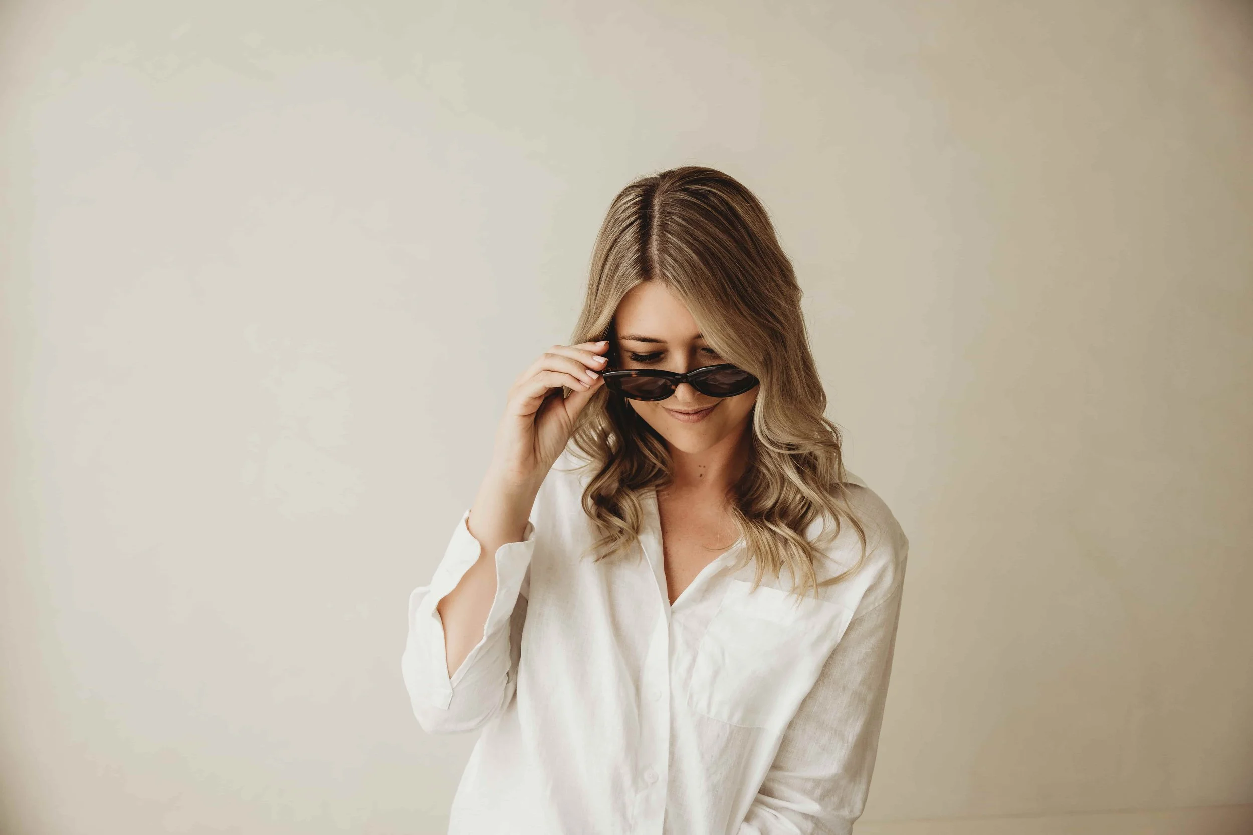 Kiara Jade Trotter wavy brown hair in a white blouse, looking down while holding black sunglasses with her right hand, against a plain beige wall.