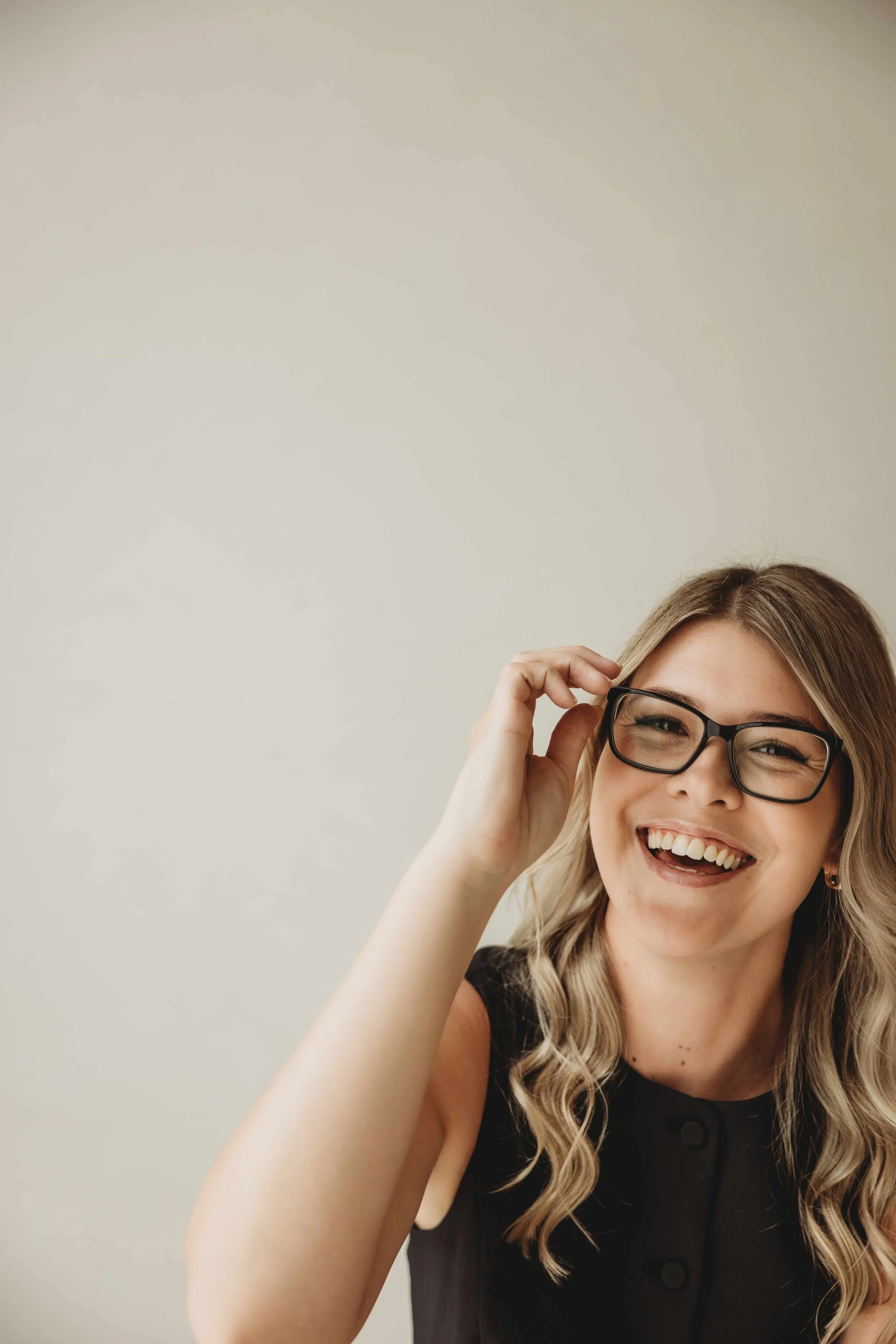 Kiara Jade Trotter with glasses smiling and adjusting her glasses with her right hand, wearing a black sleeveless top, against a plain neutral background.