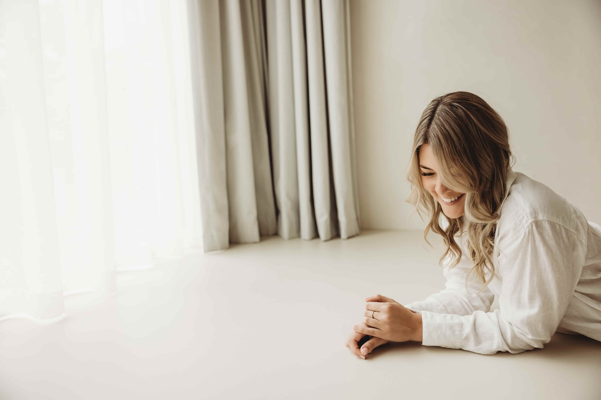 Kiara Jade Trotter with wavy blonde hair smiling while sitting on the floor near a white wall and beige curtains, with bright natural light coming through the window.