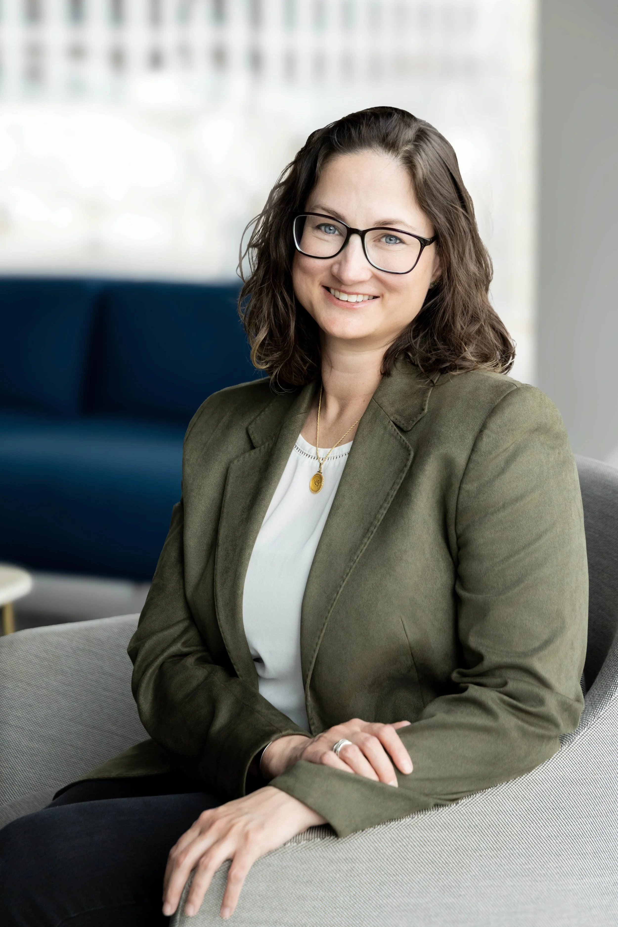 Cambium + Root Founder, Megan Mahoney, with shoulder-length brown hair, glasses, and a cheerful smile, wearing a green blazer and a white top, sitting on a beige couch in a modern, well-lit room.