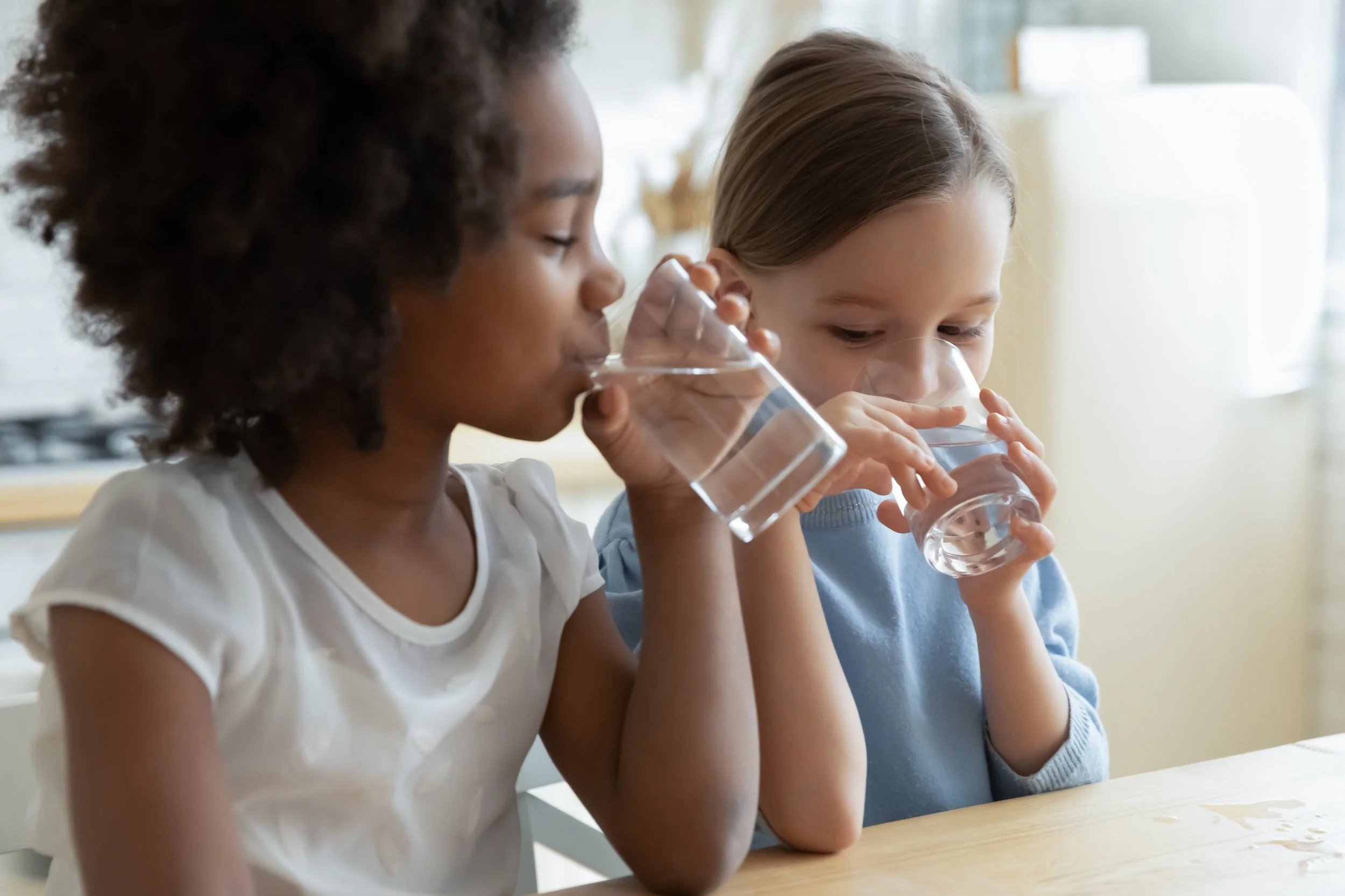 Two young girls sitting at a table drinking water from clear glasses in a bright room.