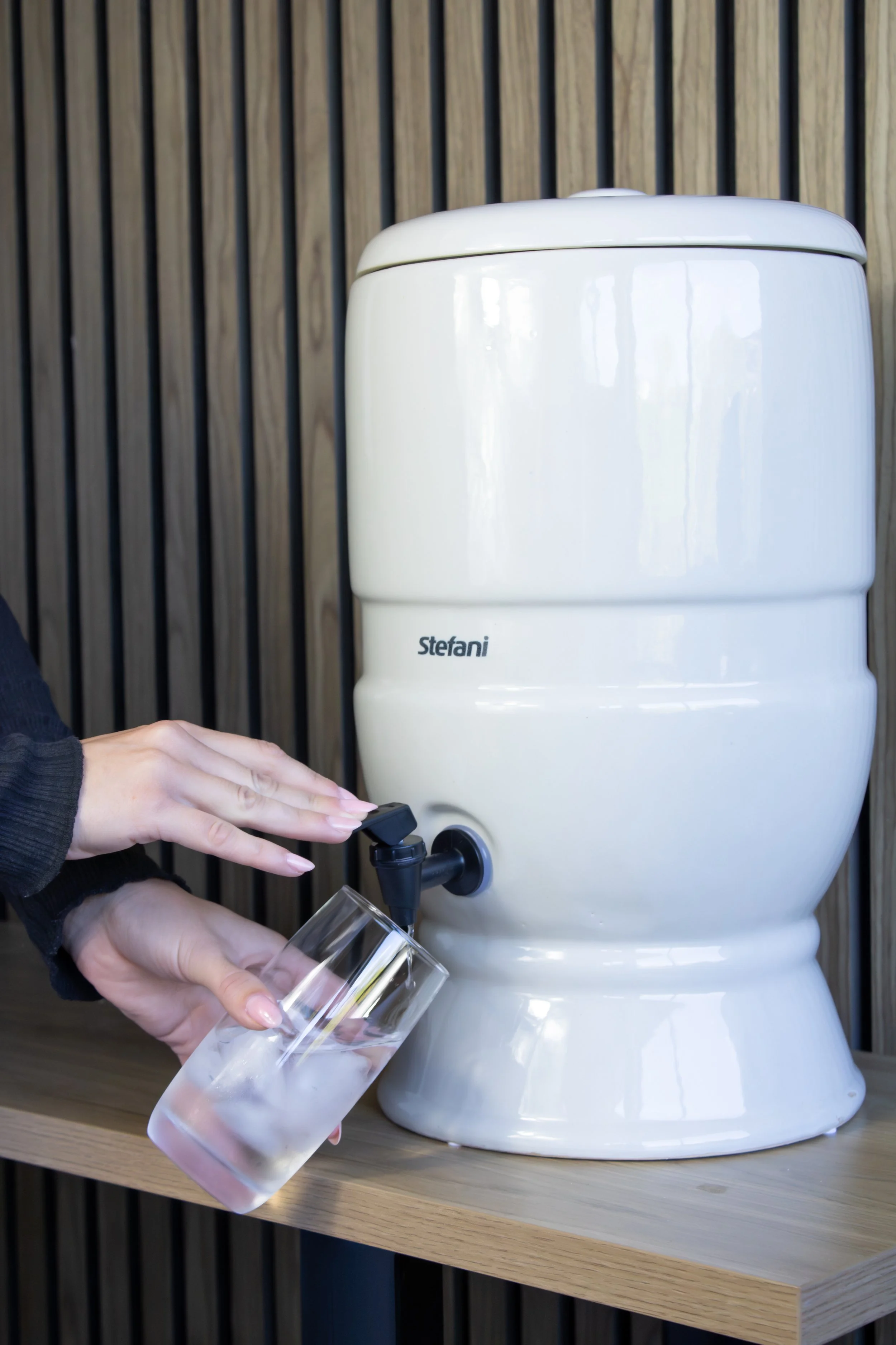 A person is filling a glass with water from a white tabletop water dispenser on a wooden surface against a wooden slat wall.
