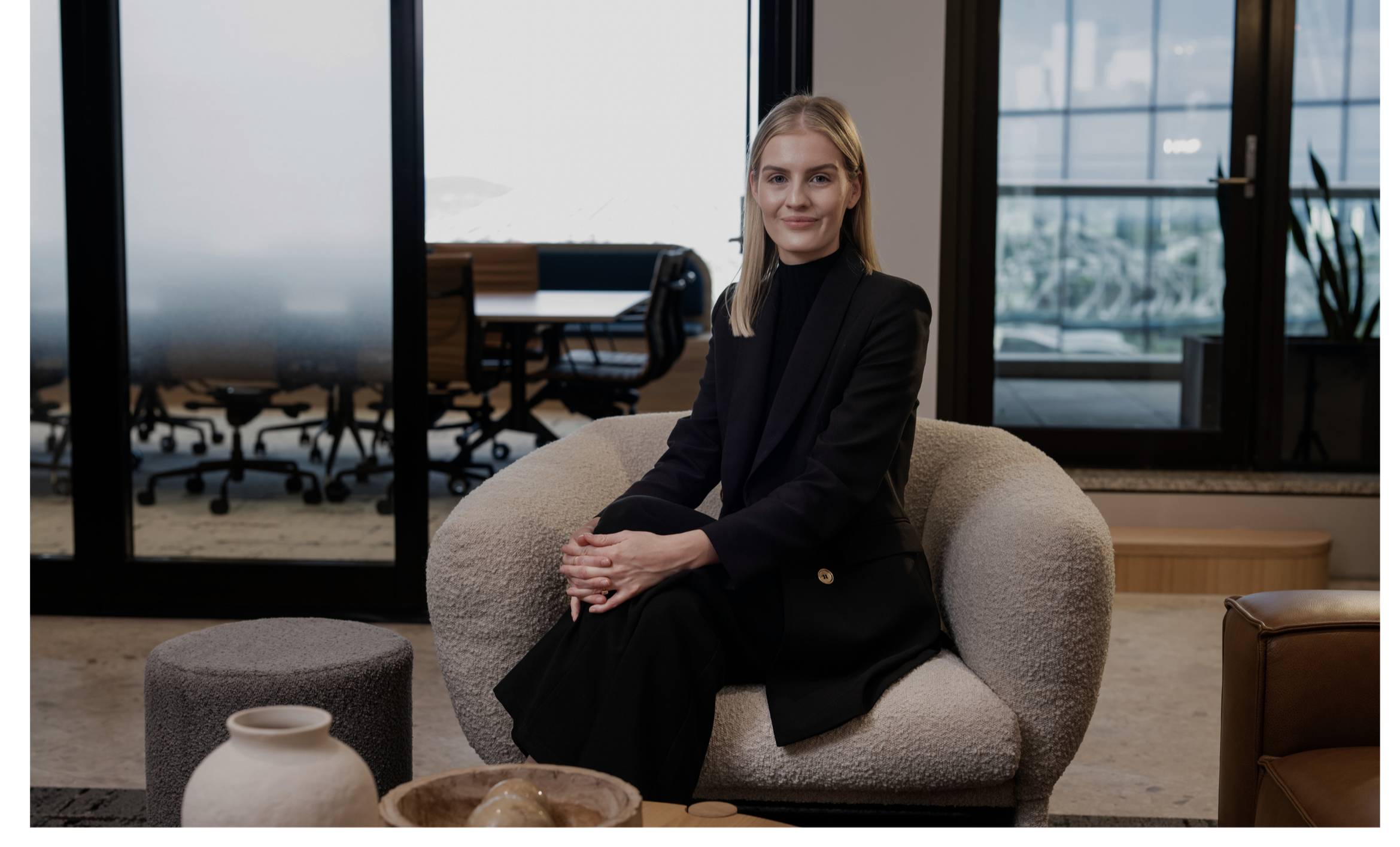 A woman with blonde hair wearing a black blazer and black dress sitting on a plush beige chair in an office lounge area with large windows and a glass door in the background.
