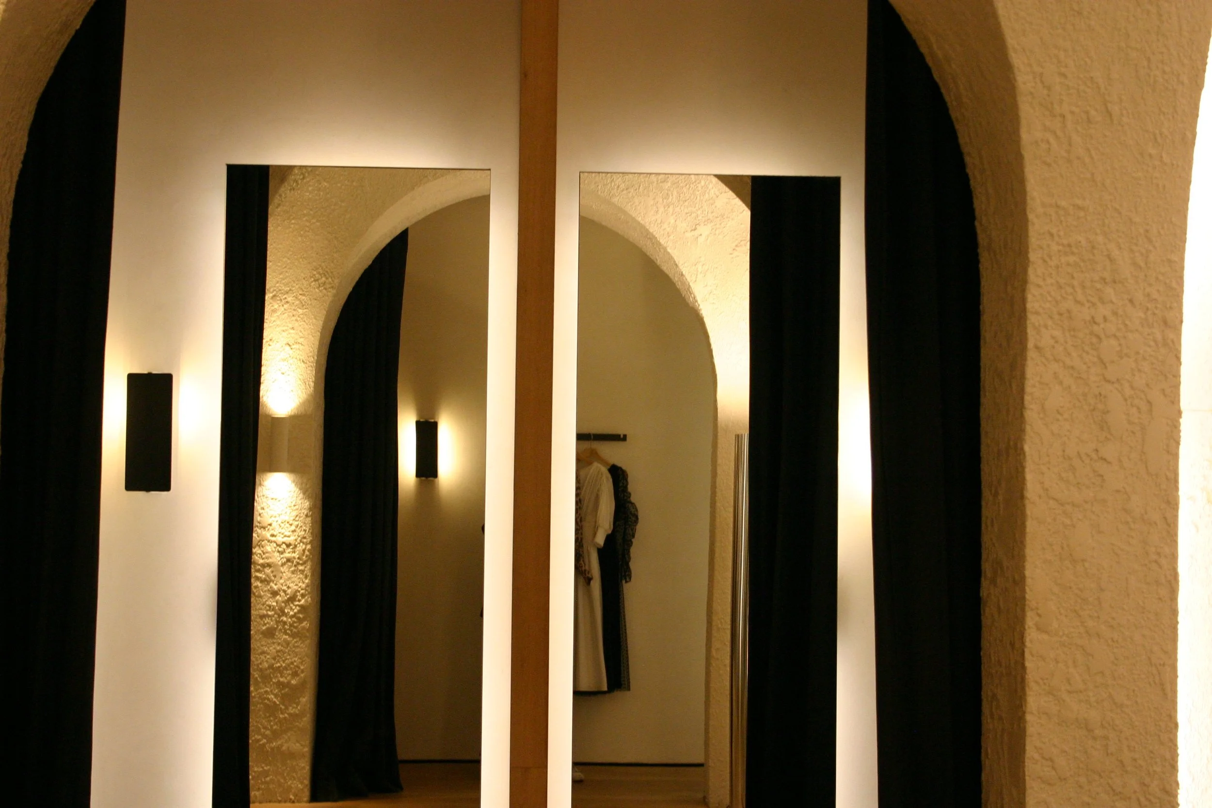 A fitting room with large mirrors, arched doorways, black curtains, textured beige walls, and clothing on a rack in the background.