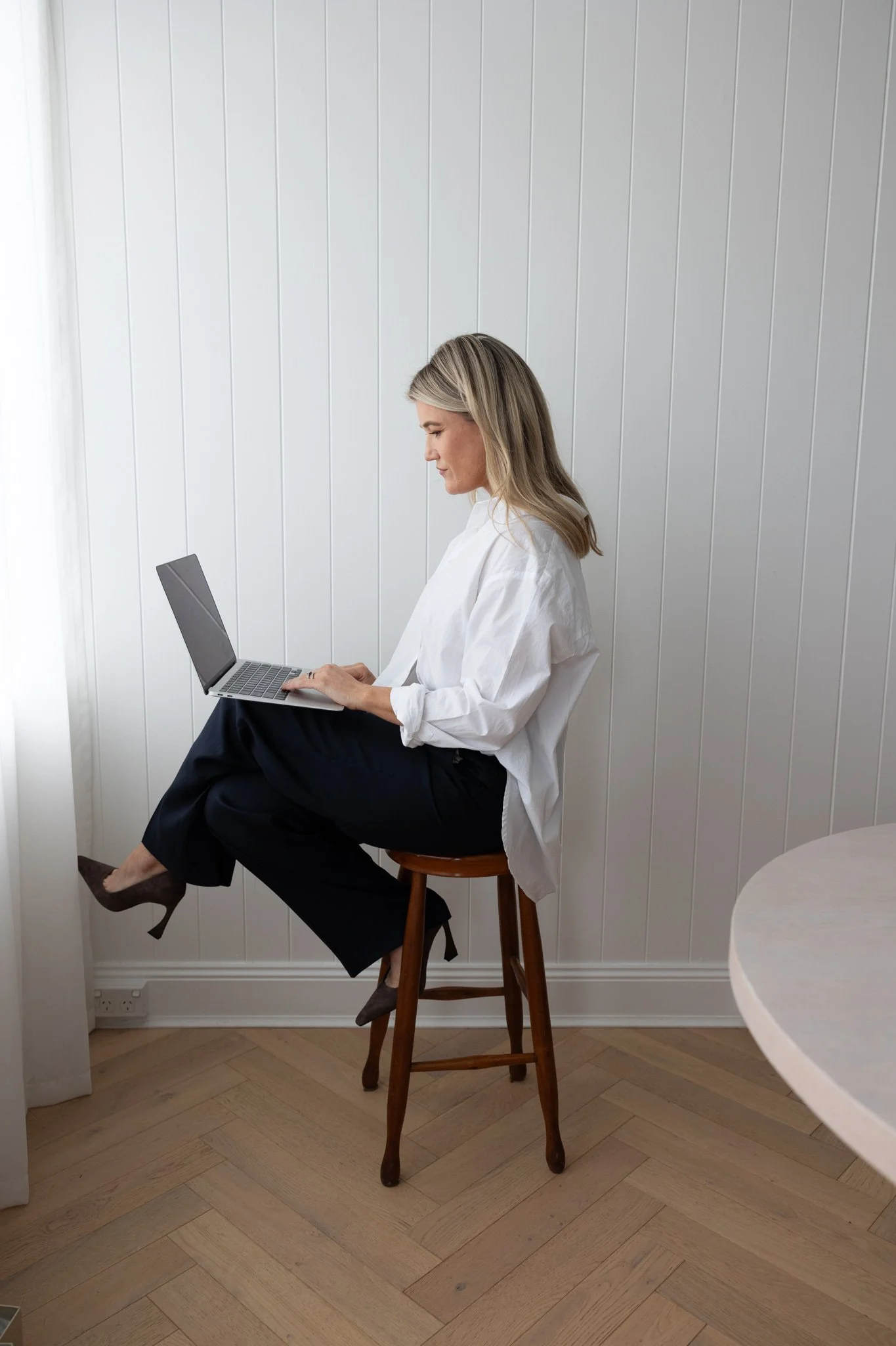 woman sitting on a wooden stool using a laptop in a room with white paneled walls and wooden flooring.