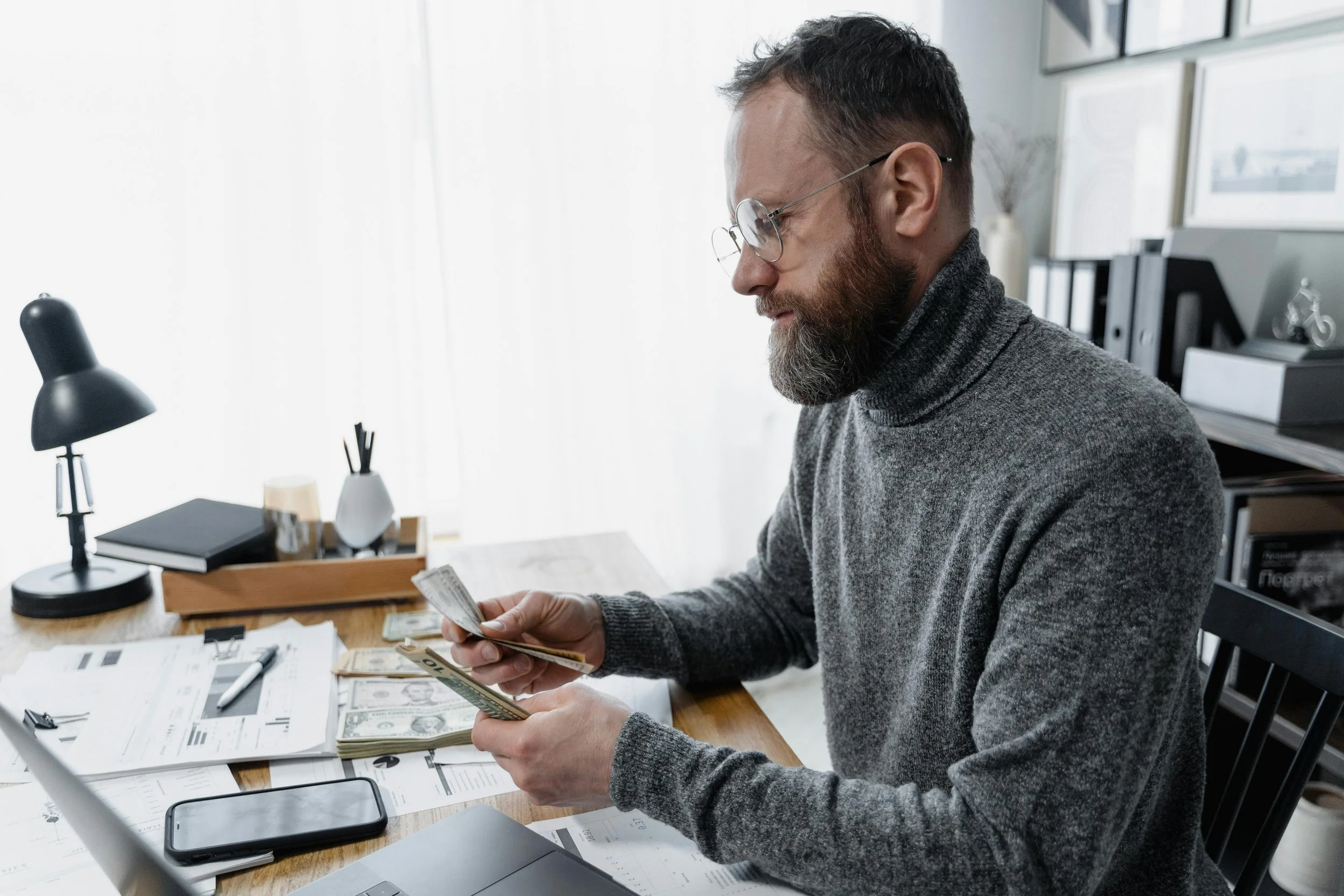 Man counting cash at a desk with a laptop, smartphone, and paperwork. Represents capital sourcing and financial planning for EB5 visa investments and the path to U.S. permanent residency through the immigrant investor program.