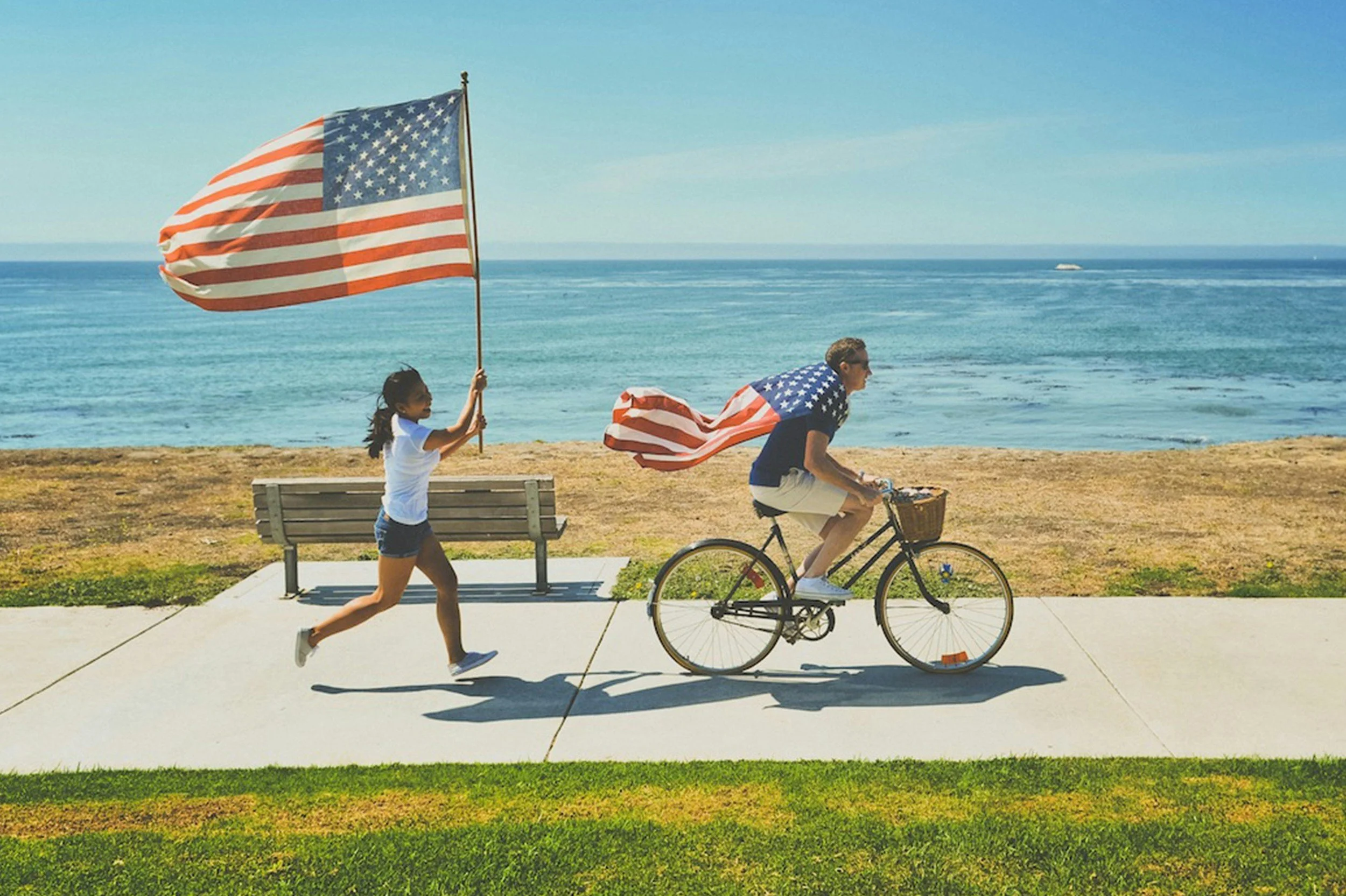 A young couple celebrates life in the U.S. as new EB5 Green Card holders—one riding a bike draped in the American flag, the other joyfully running with a large flag along a coastal path.
