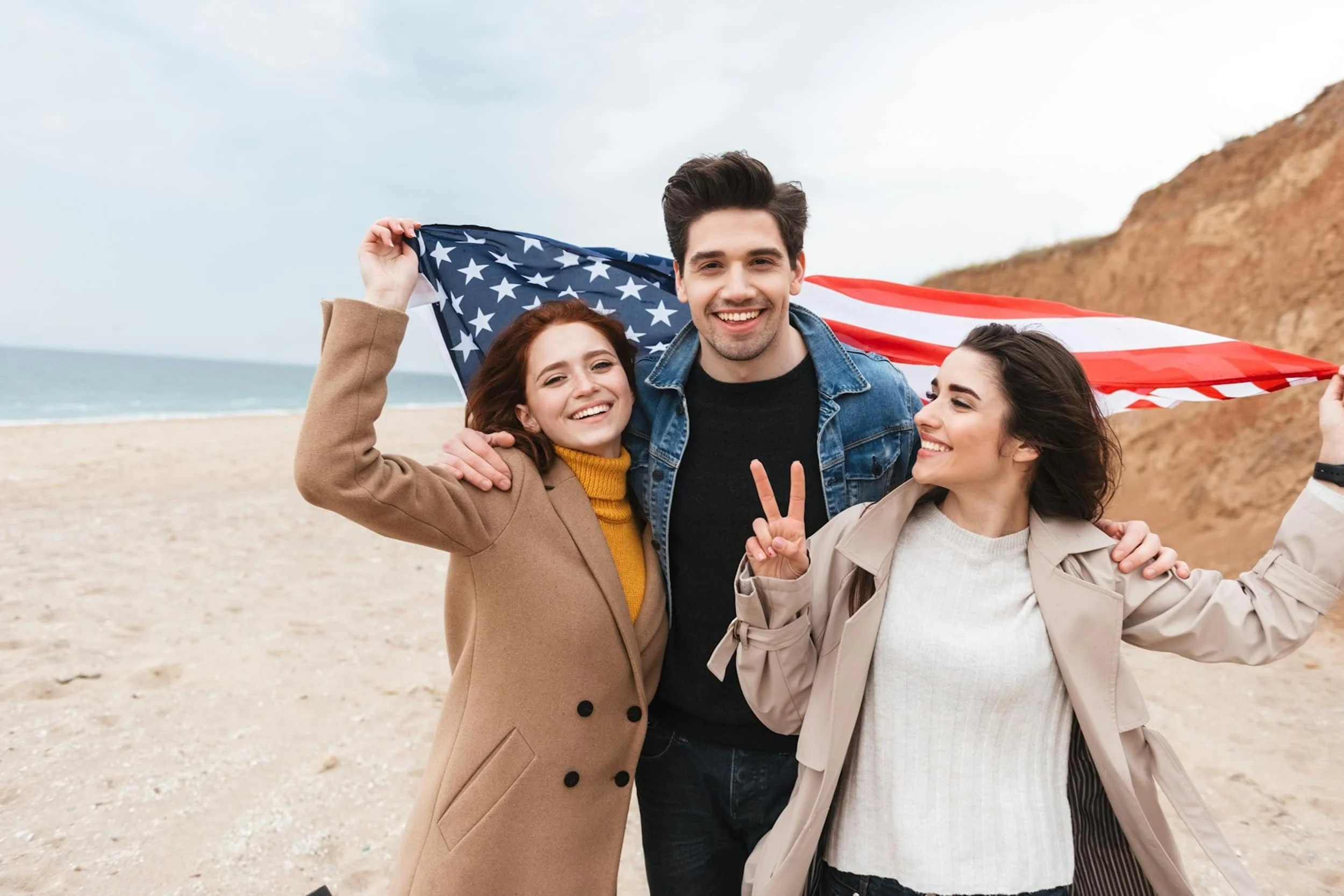 Three happy immigrants holding an American flag on a beach, celebrating EB5 Green Card approval and a new life in the United States with the help of Arcasia Advisors.