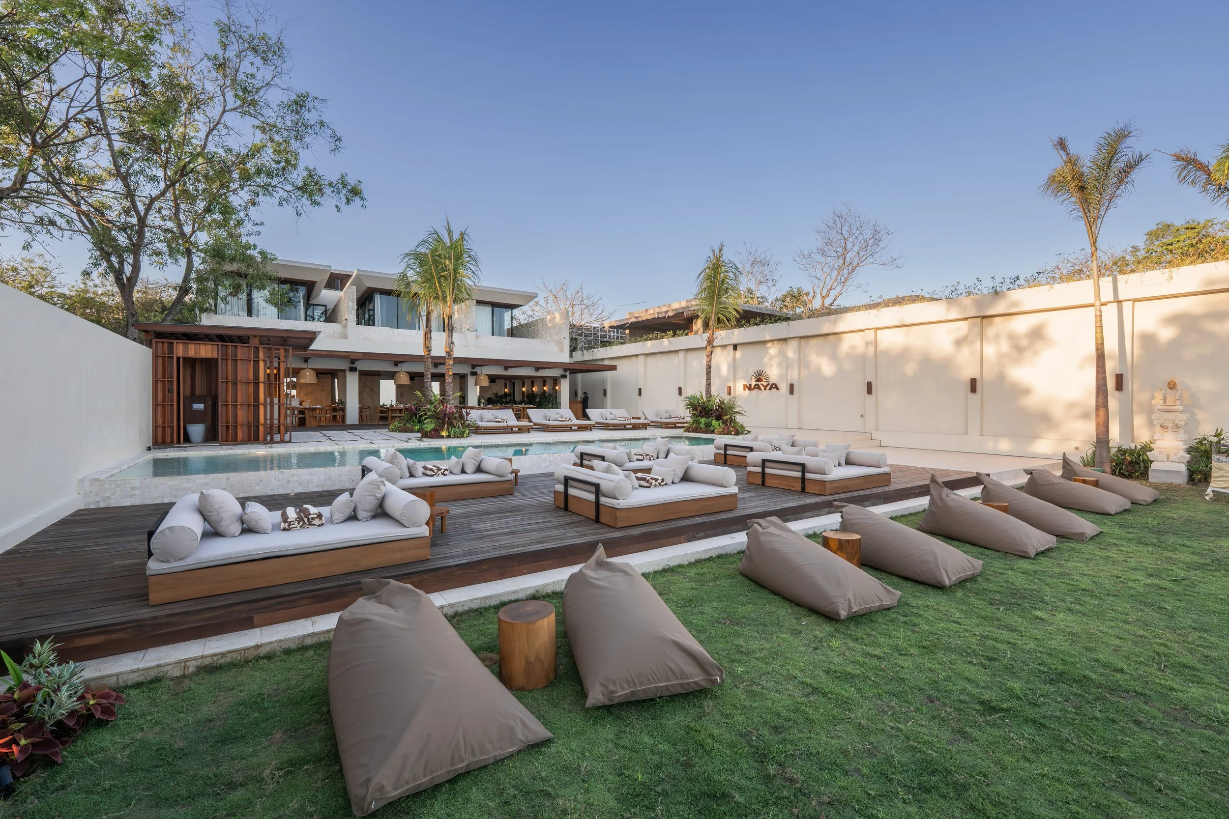 An outdoor pool area with lounge chairs, bean bag seats, and palm trees, adjacent to a modern, multi-story building with large glass windows and a restaurant or bar with hanging lights.