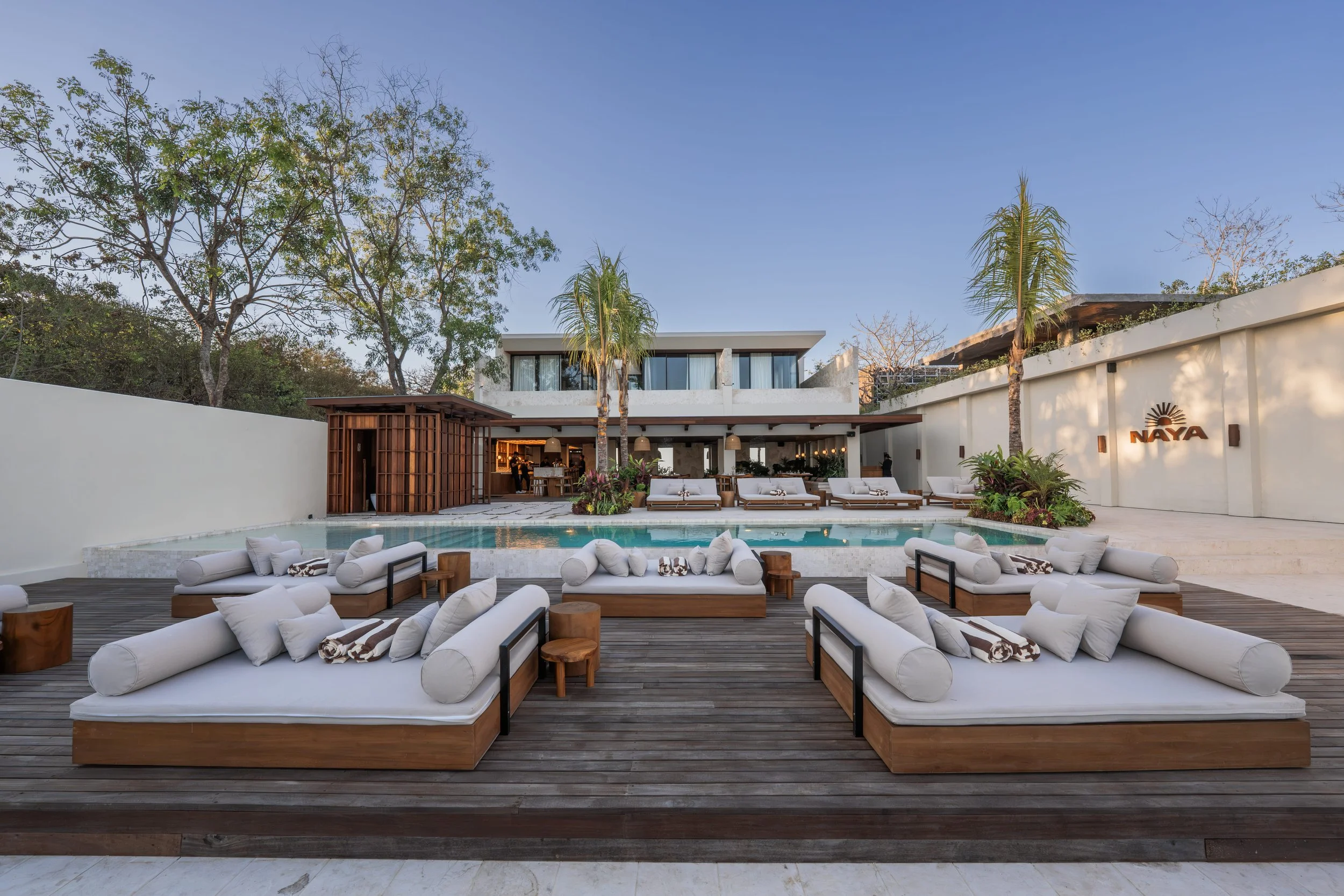 Luxurious outdoor poolside lounge area with white cushioned daybeds and small wooden tables, modern building with large windows in the background, lush green trees, and clear blue sky.