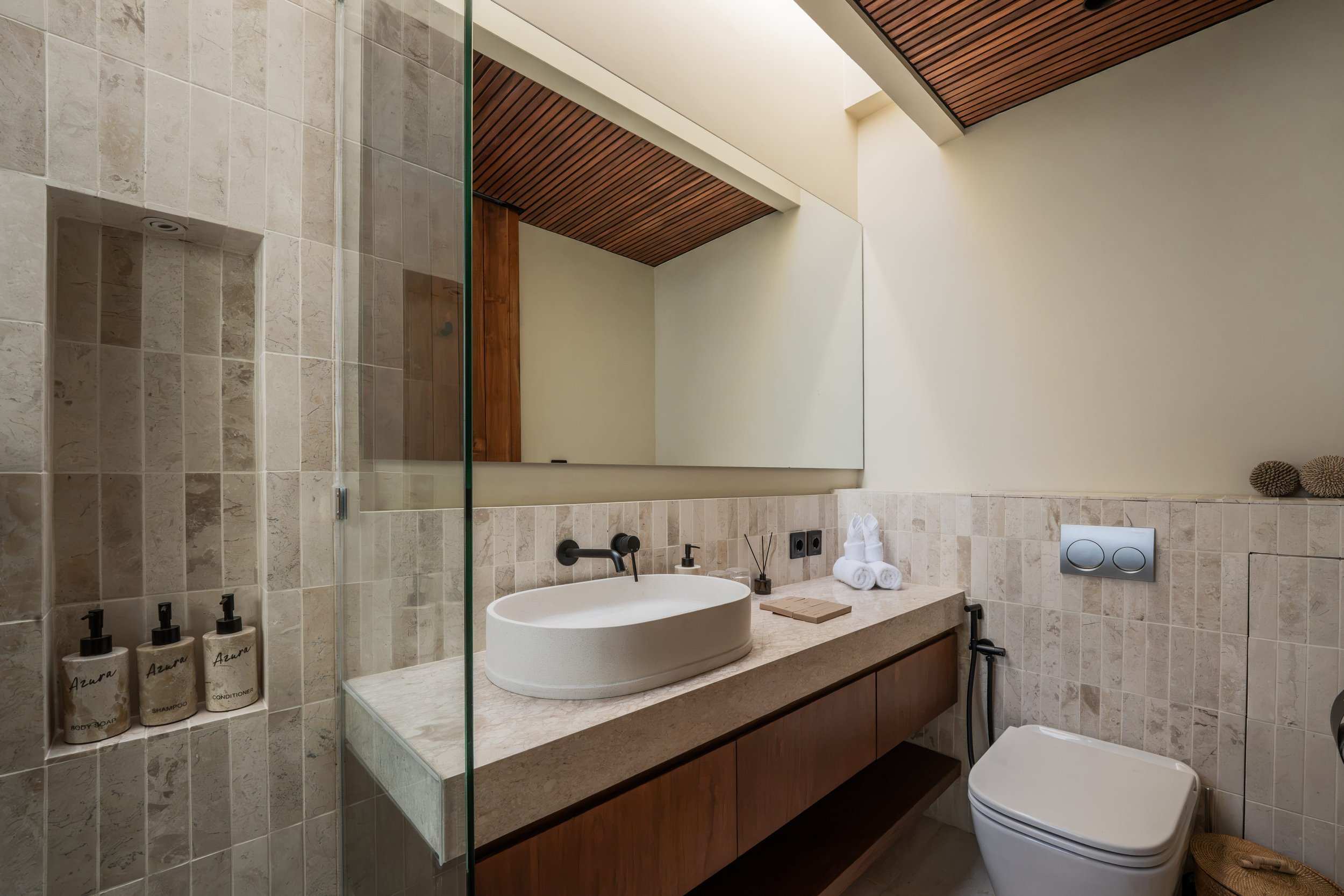 Modern bathroom with beige tiled walls, a white vessel sink, black faucet, and wooden cabinetry below. Large mirror above the sink, decorated with rolled towels, toiletries, and a small tray. A glass shower enclosure with built-in niche for toiletrie