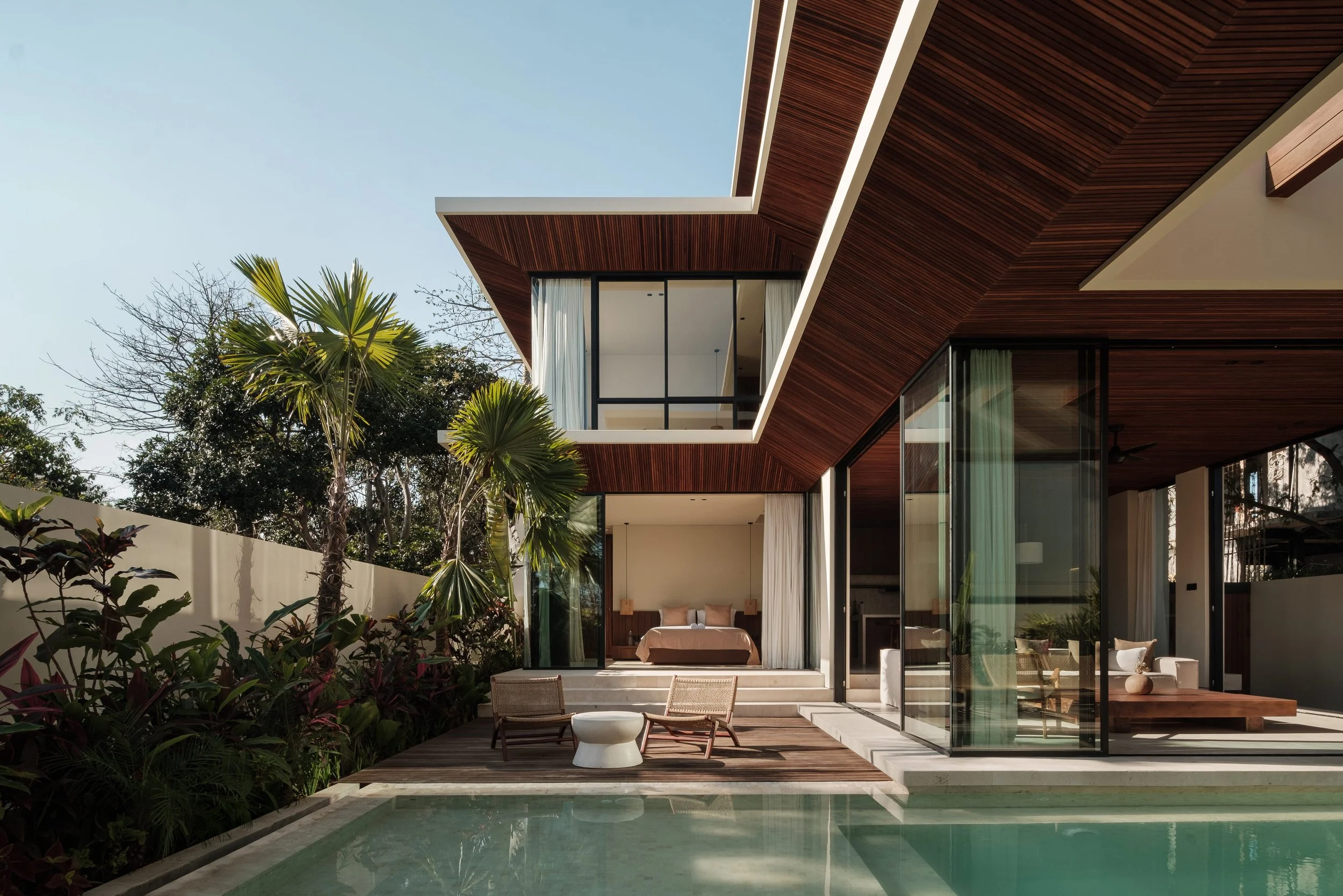 Modern two-story house with extensive glass windows, wood paneling, patio furniture, and a swimming pool surrounded by lush tropical plants.