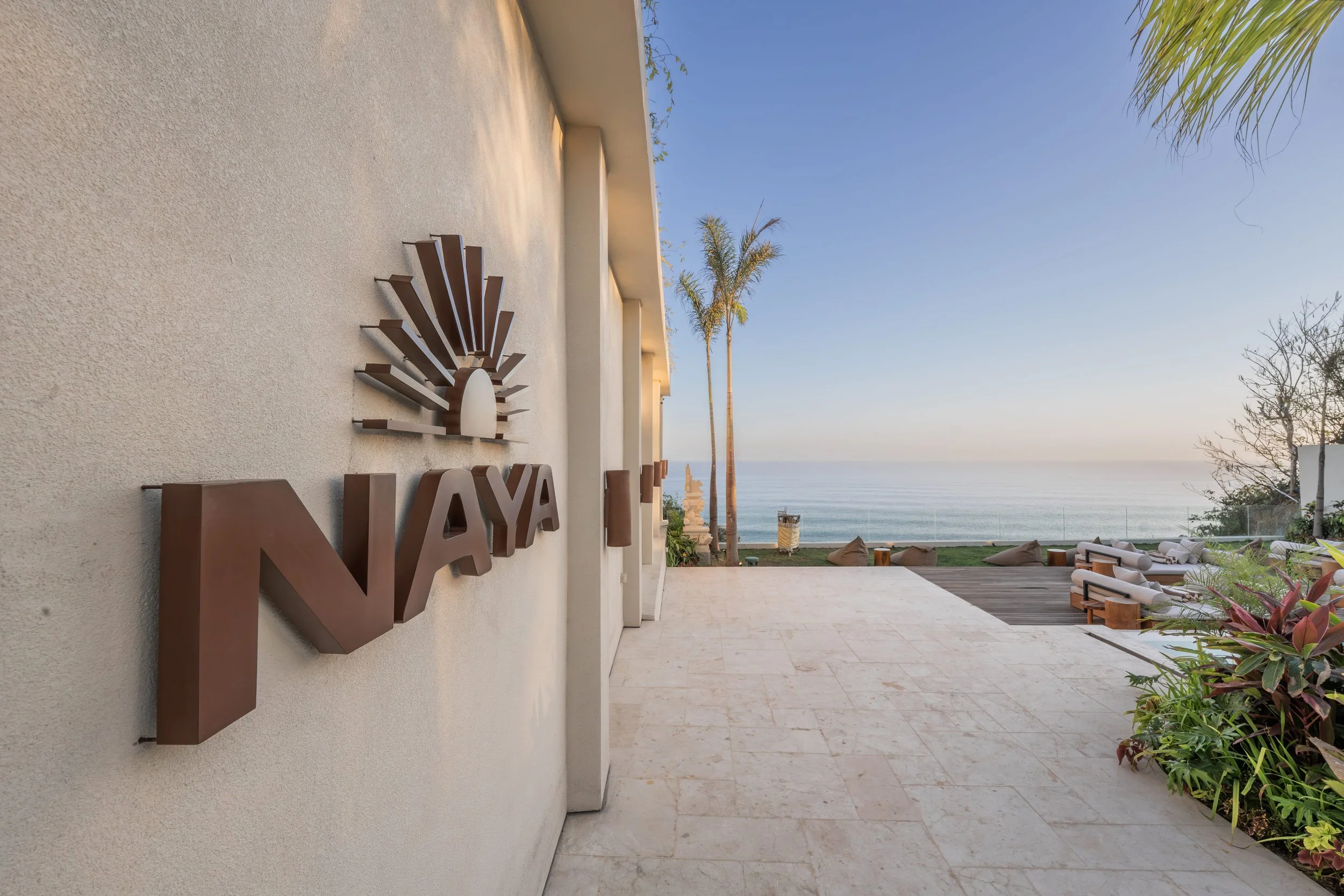 A view of a modern outdoor terrace with a beige wall featuring a metal sunburst logo and the letters 'NAYA.' The terrace overlooks the ocean and contains palm trees, lounge chairs, and plants, under a clear blue sky.