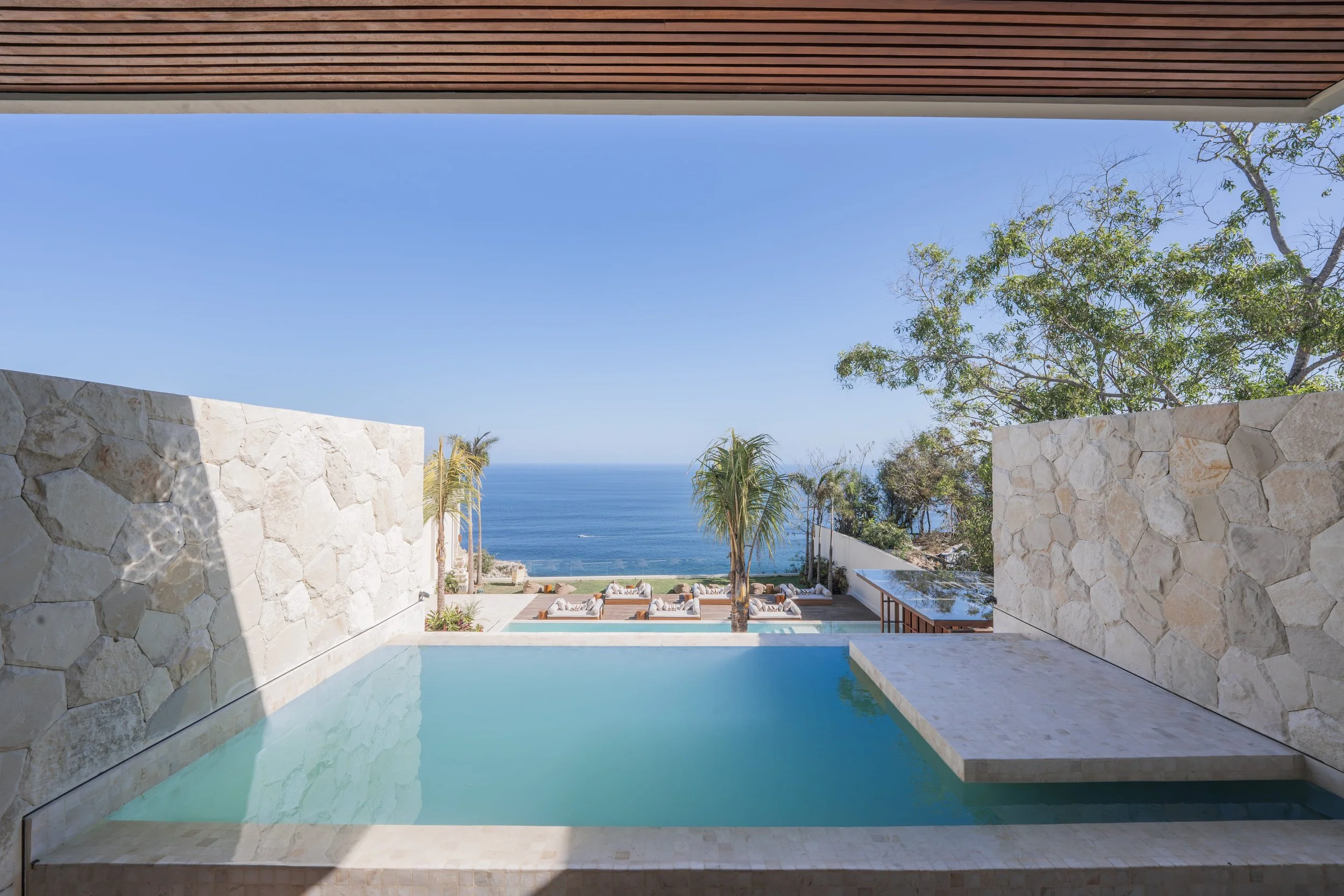View of a modern outdoor pool area with white stone walls and lounge chairs, overlooking the ocean with trees and blue sky in the background.