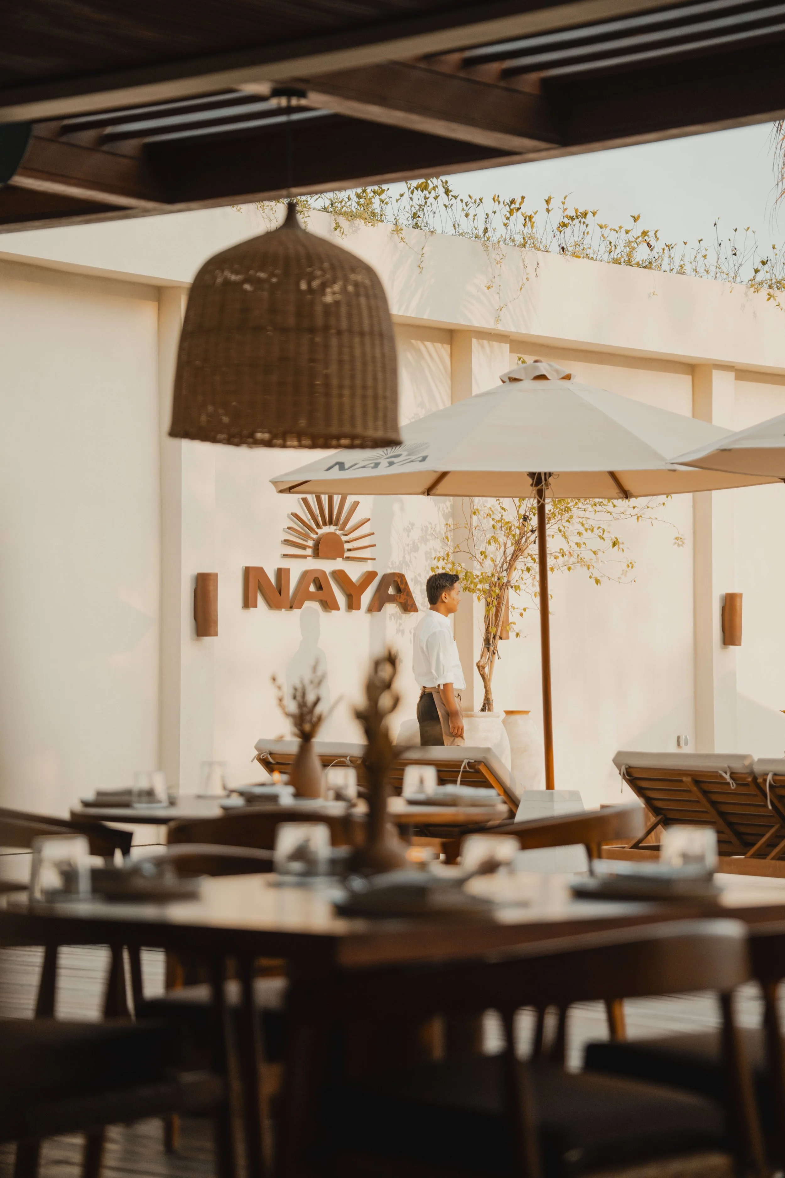 A restaurant or cafe with tables and chairs in the foreground, a person walking past a wall with the word 'NAYA' on it, patio umbrellas, and outdoor greenery visible.