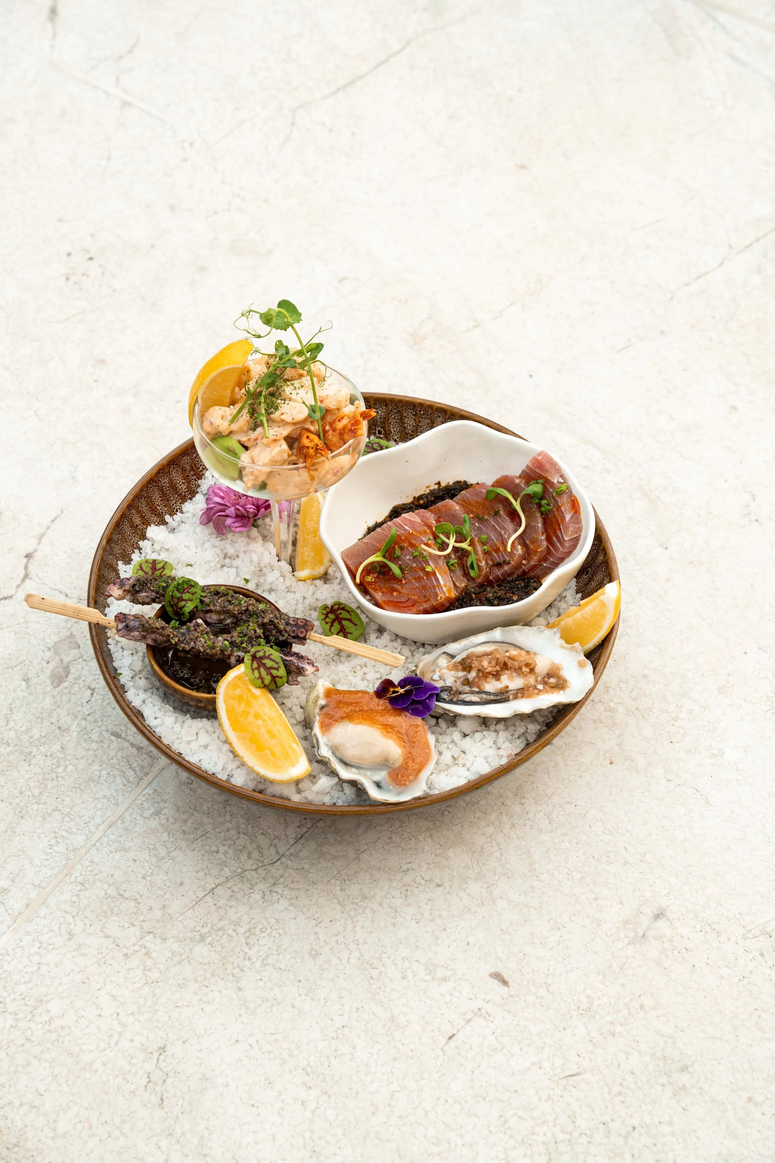 A large oval serving platter filled with raw seafood and garnishes, including oysters, slices of lemon, slices of sashimi, shrimp, and a cocktail shrimp in a small glass with garnishes on a light surface.