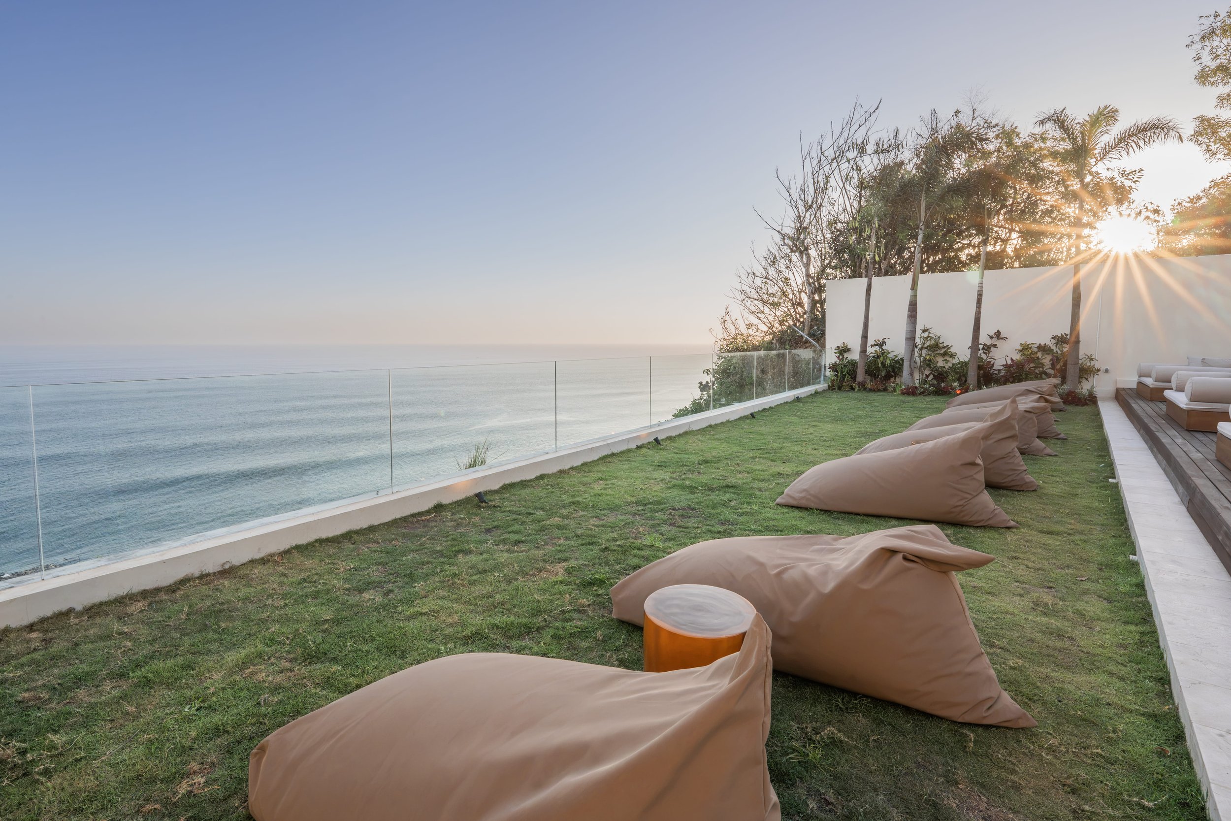 Outdoor rooftop terrace with bean bag chairs, small tables, a grassy area, and a clear glass railing overlooking the ocean at sunset.