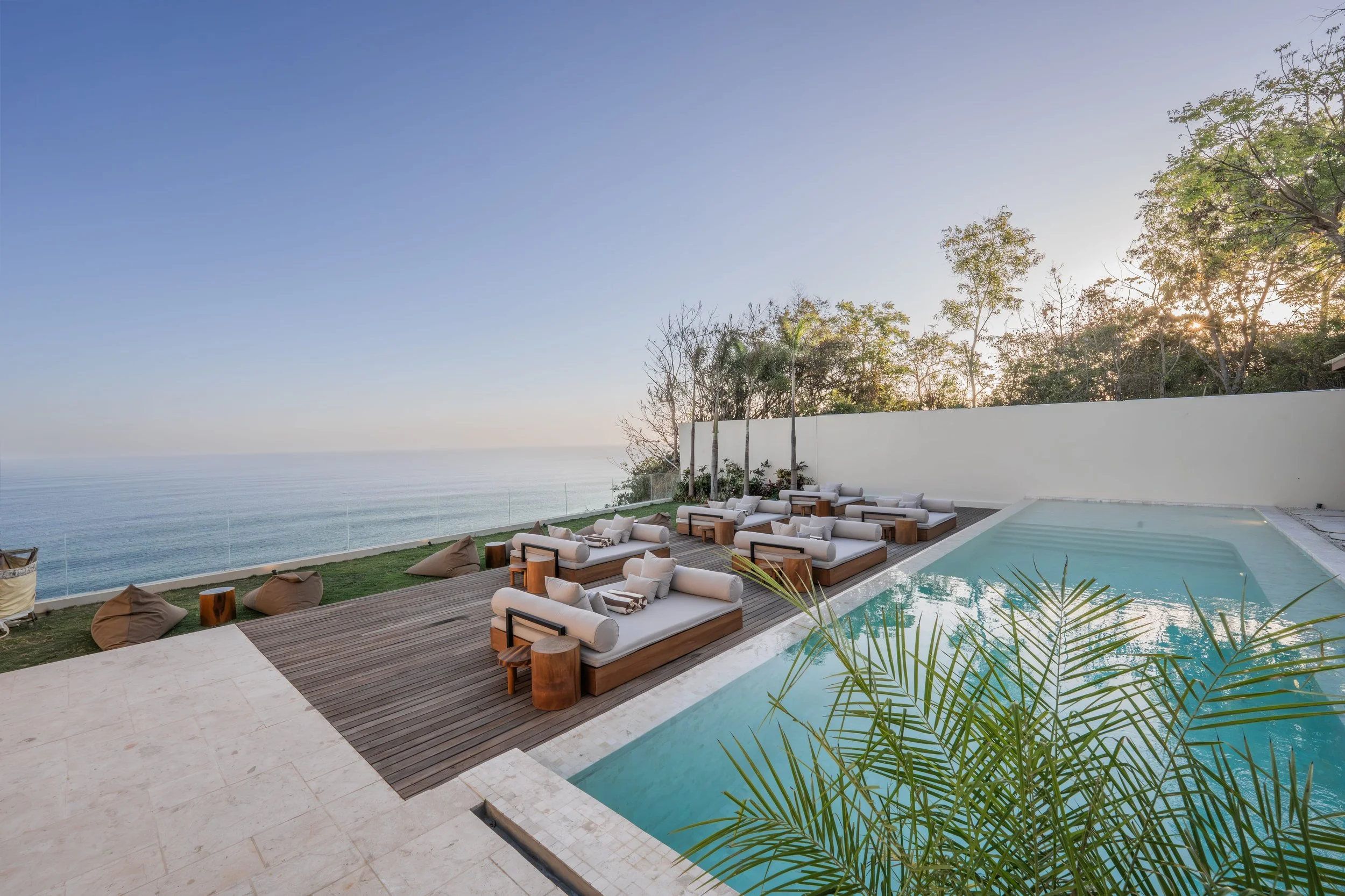 Luxury outdoor pool area overlooking the ocean with multiple lounge chairs, bean bags, and a white wall with trees behind it, under a clear sky.