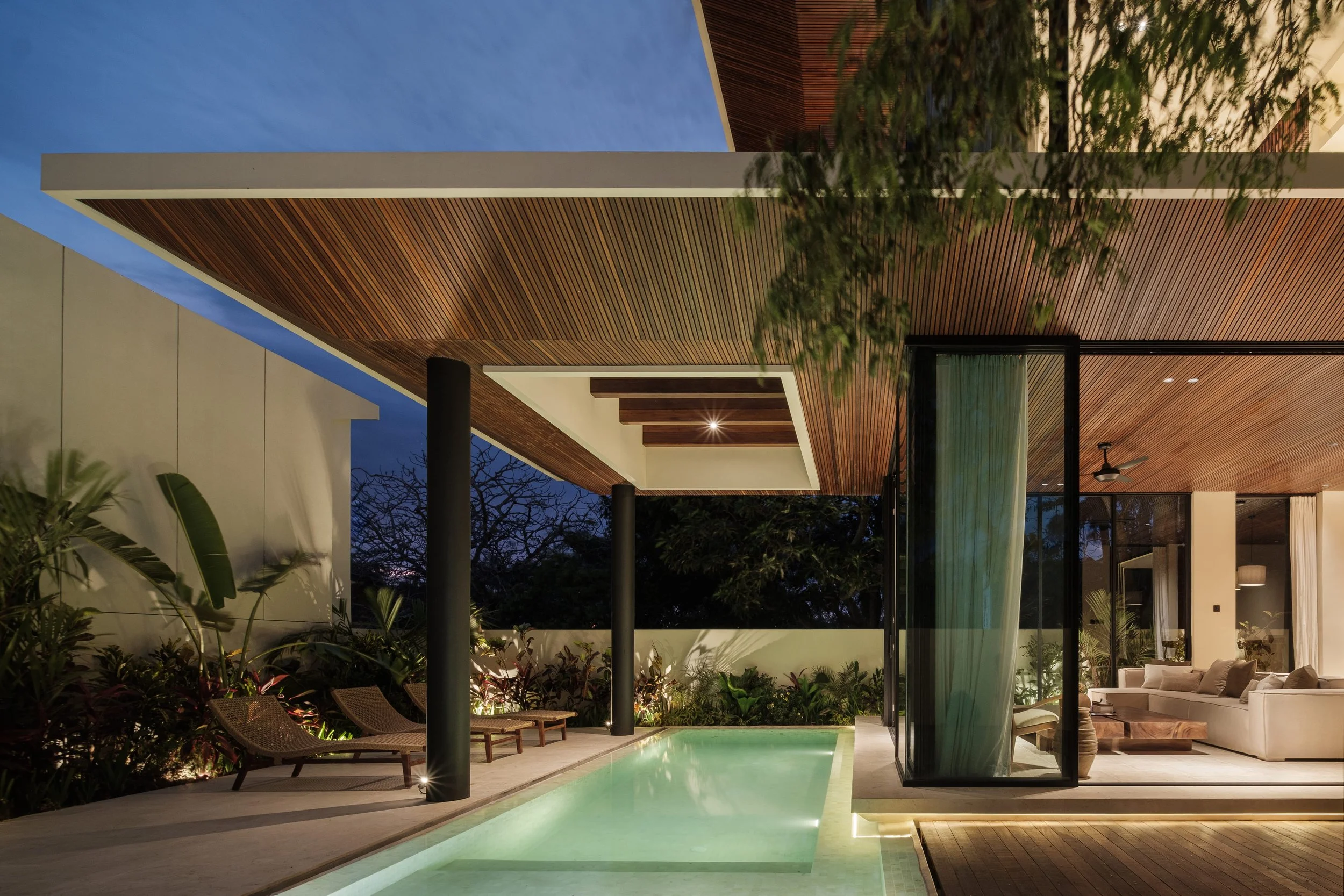 Modern house exterior at night with a swimming pool, wooden ceiling, and outdoor furniture, illuminated by ambient lighting.