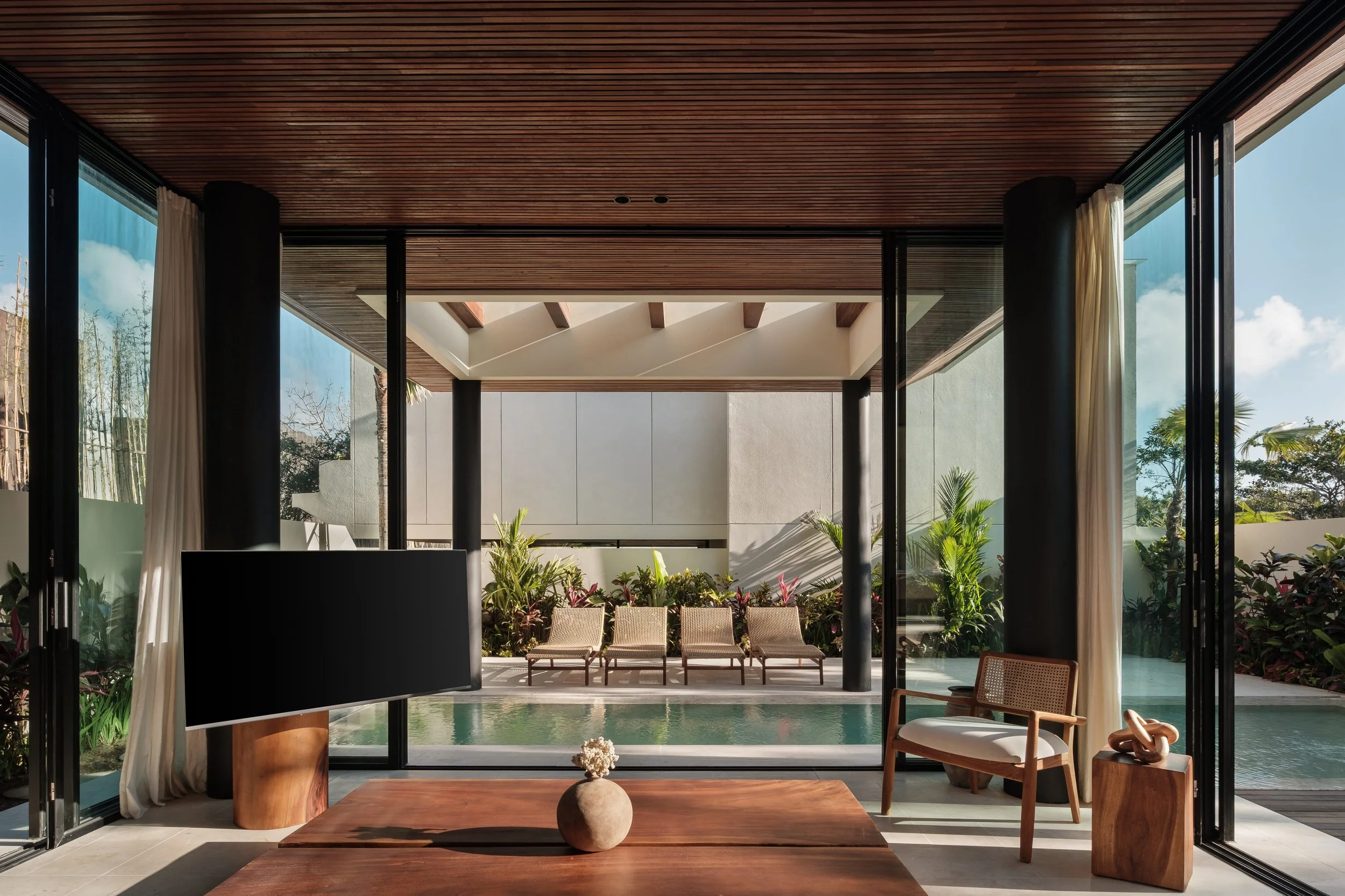 Interior of a modern house with large open sliding glass doors, wooden ceiling, and outdoor patio with chairs, plants, and a view of trees.