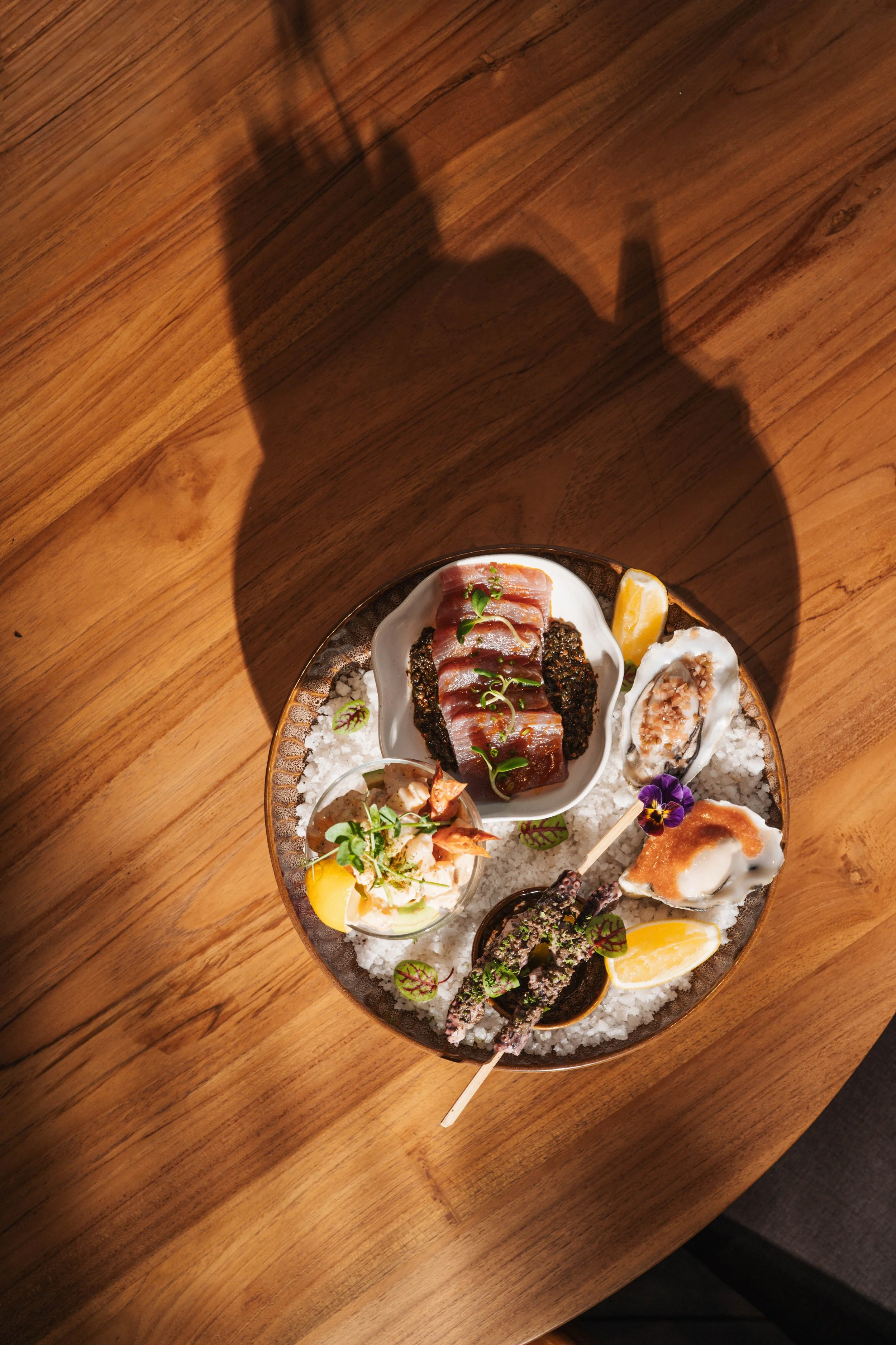 A round platter of assorted seafood dishes on a wooden table, including oysters, sashimi, garnishes, and lemon wedges.