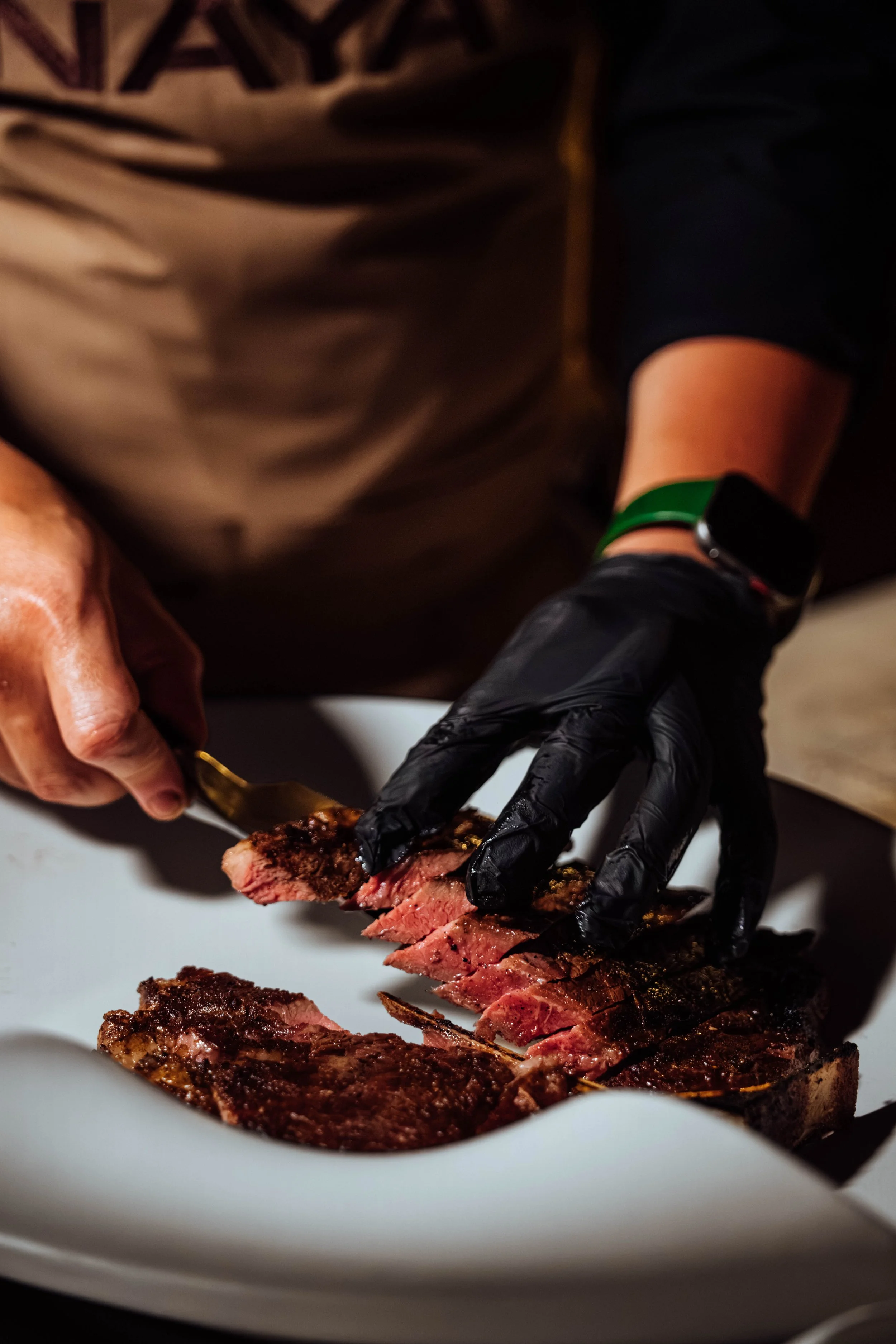 A person wearing black gloves and a fitness tracker, slicing a cooked steak on a white plate.