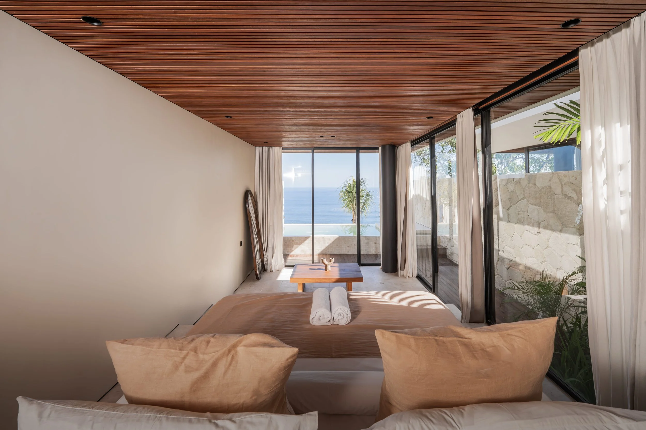 Interior view of a modern bedroom with a wooden ceiling, large glass sliding doors with curtains, and a view of the ocean and palm trees outside.