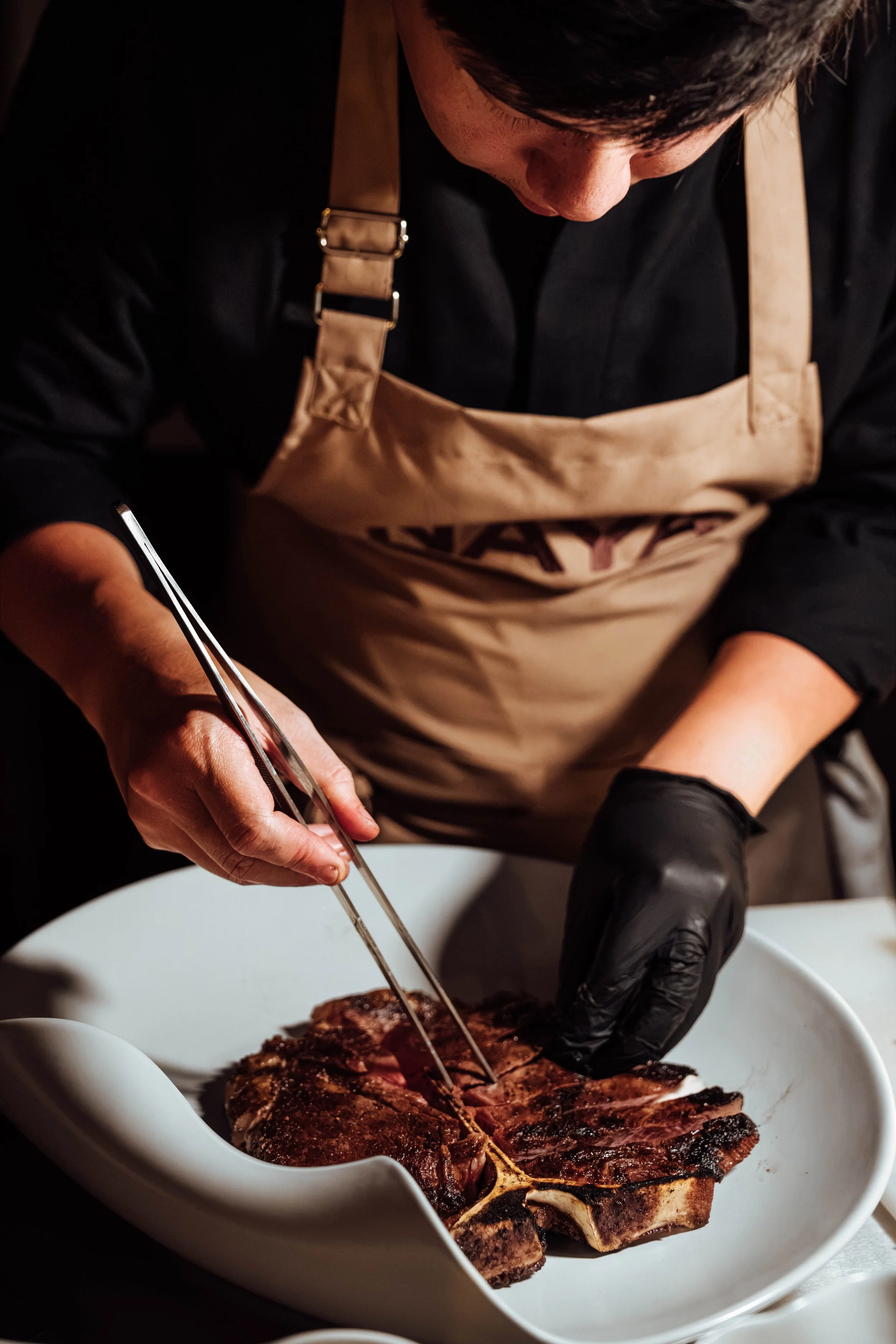 Person in black shirt and tan apron carving cooked meat in a white dish.