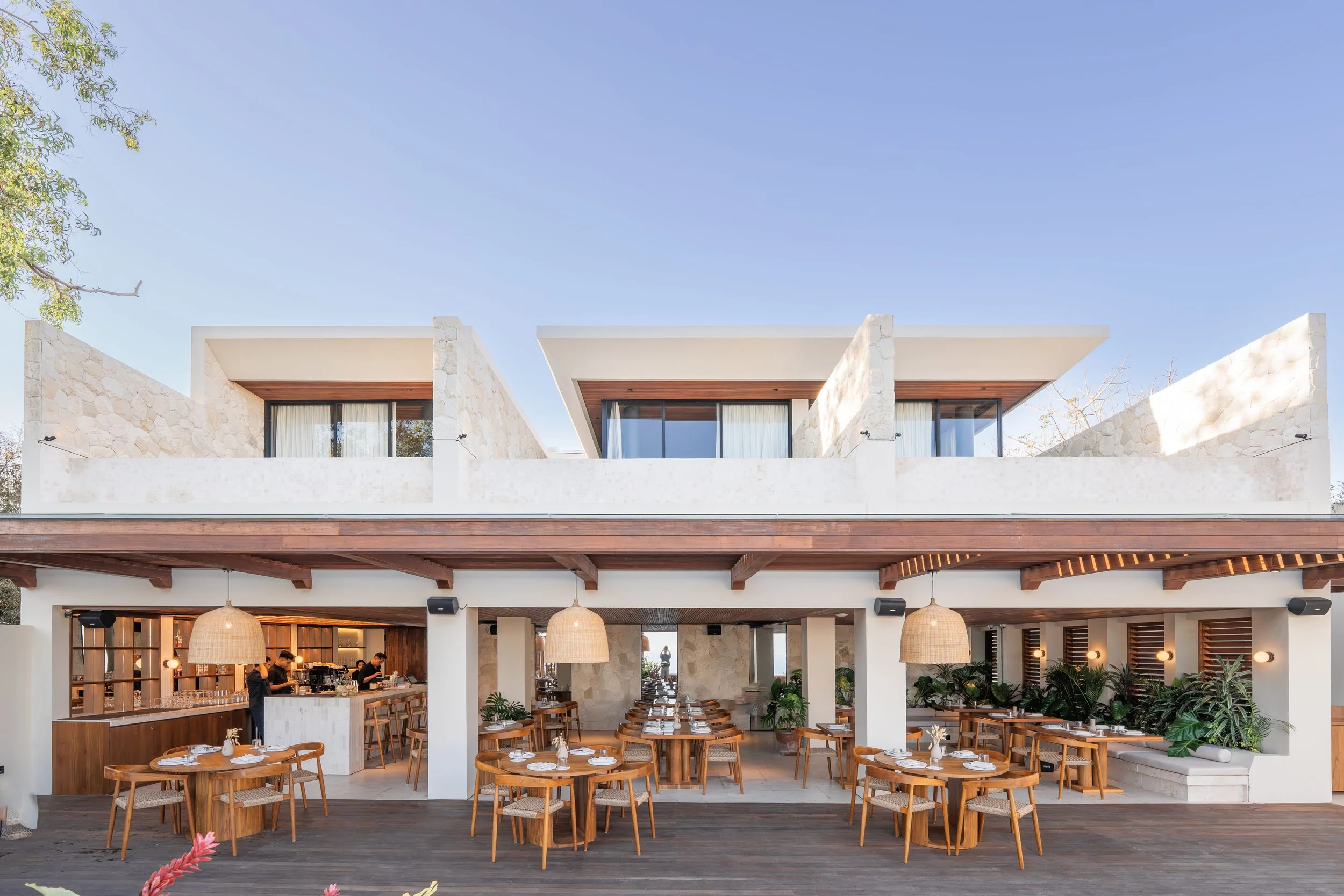 The image shows the interior of a modern, stylish restaurant or cafe with wooden tables and chairs, some set for dining with plates, napkins, and small decorations. The space features a high ceiling with hanging woven light fixtures, large windows, a