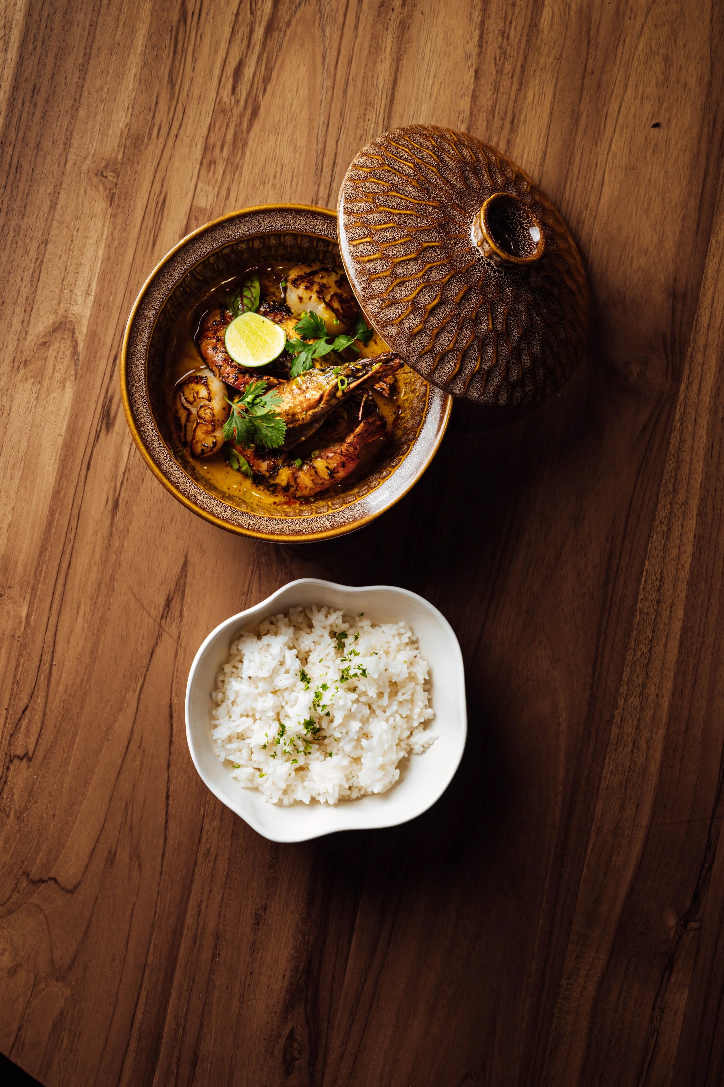 A bowl of Asian-style seafood curry with shrimp, scallops, lime, and herbs, alongside a bowl of white rice on a wooden table.