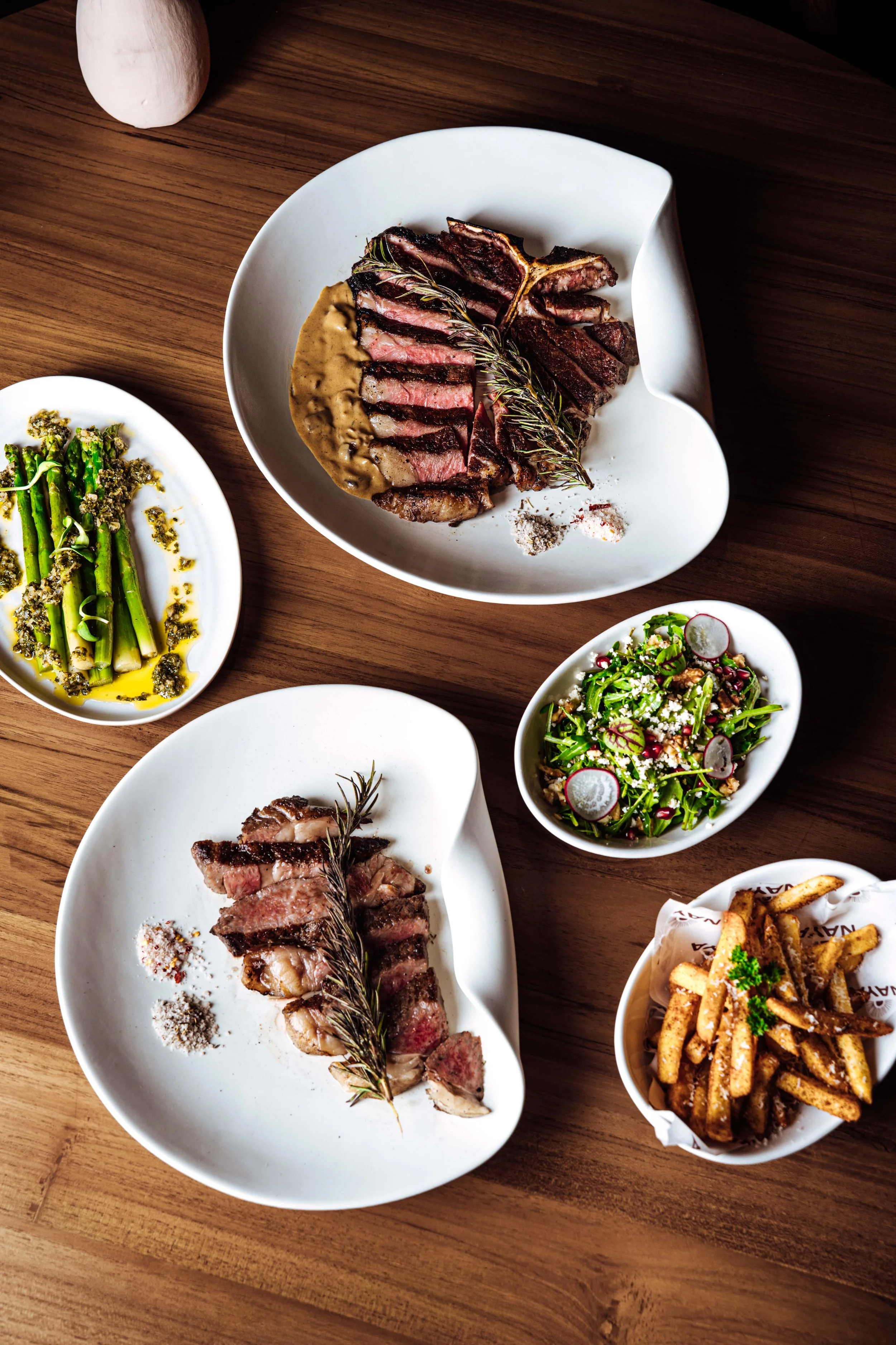 Assorted dishes on a wooden table including sliced rare steak with a sprig of rosemary and sauce, green asparagus with pesto, mixed vegetable salad with radishes, and a bowl of seasoned French fries.