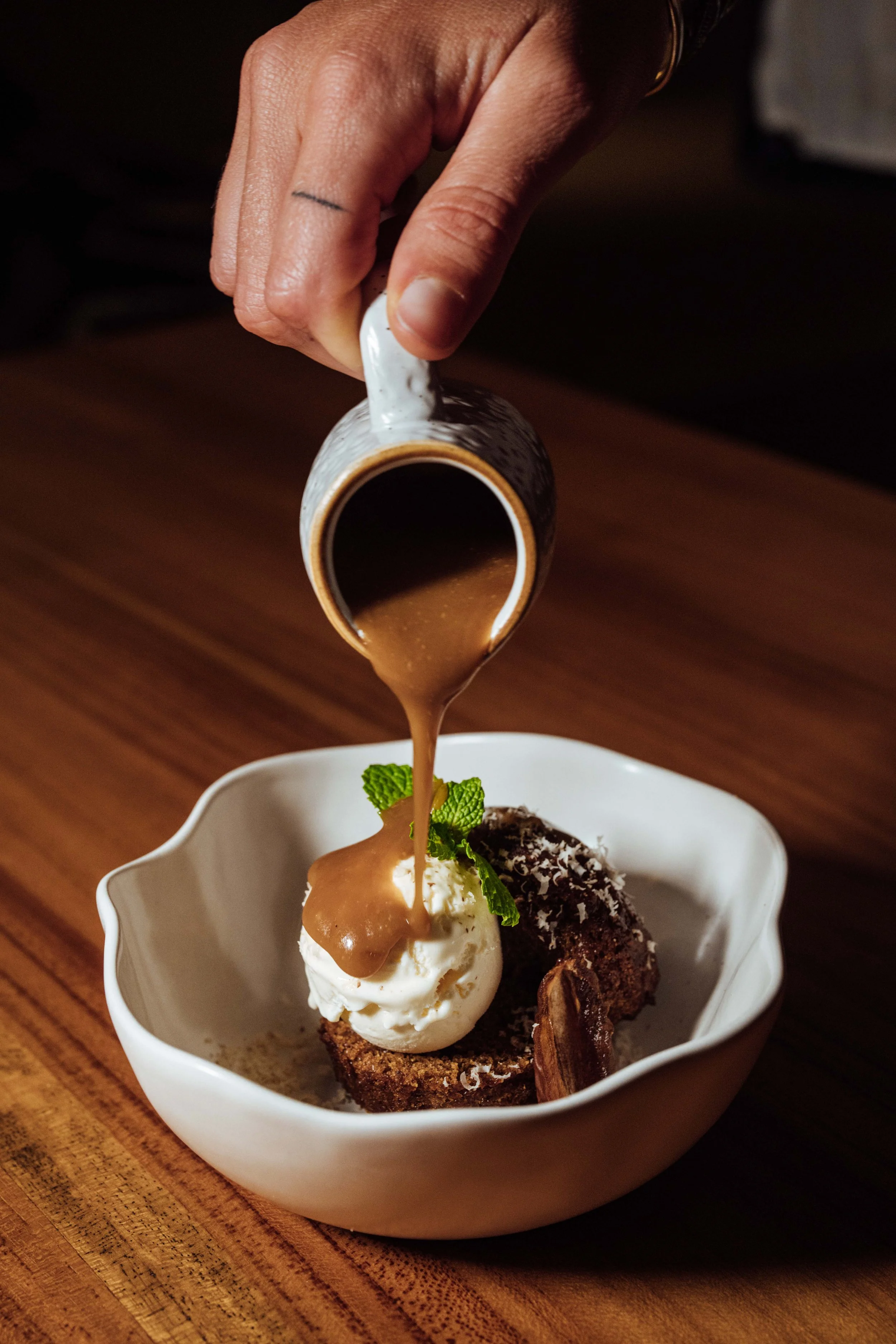 A small white bowl holding a bowl of ice cream with a brownie, whipped cream, mint leaves, and caramel sauce. A hand pours caramel sauce from a patterned mug over the ice cream.