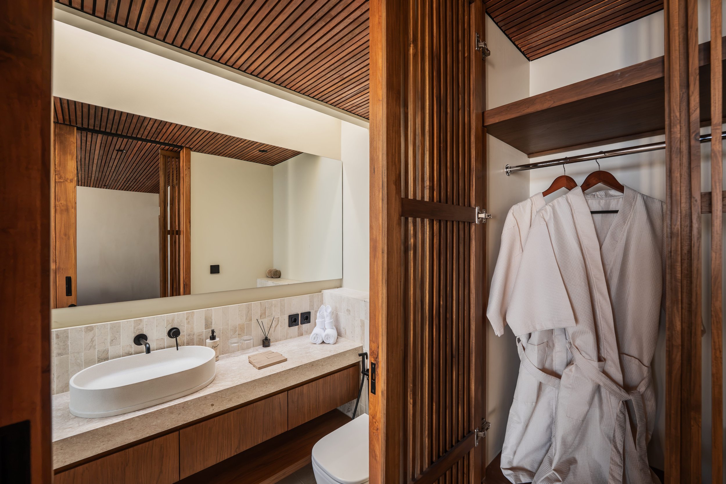 Modern bathroom with wooden accents, a large mirror, a white countertop with a vessel sink, toiletries, and a robe hanging on a wooden closet door.