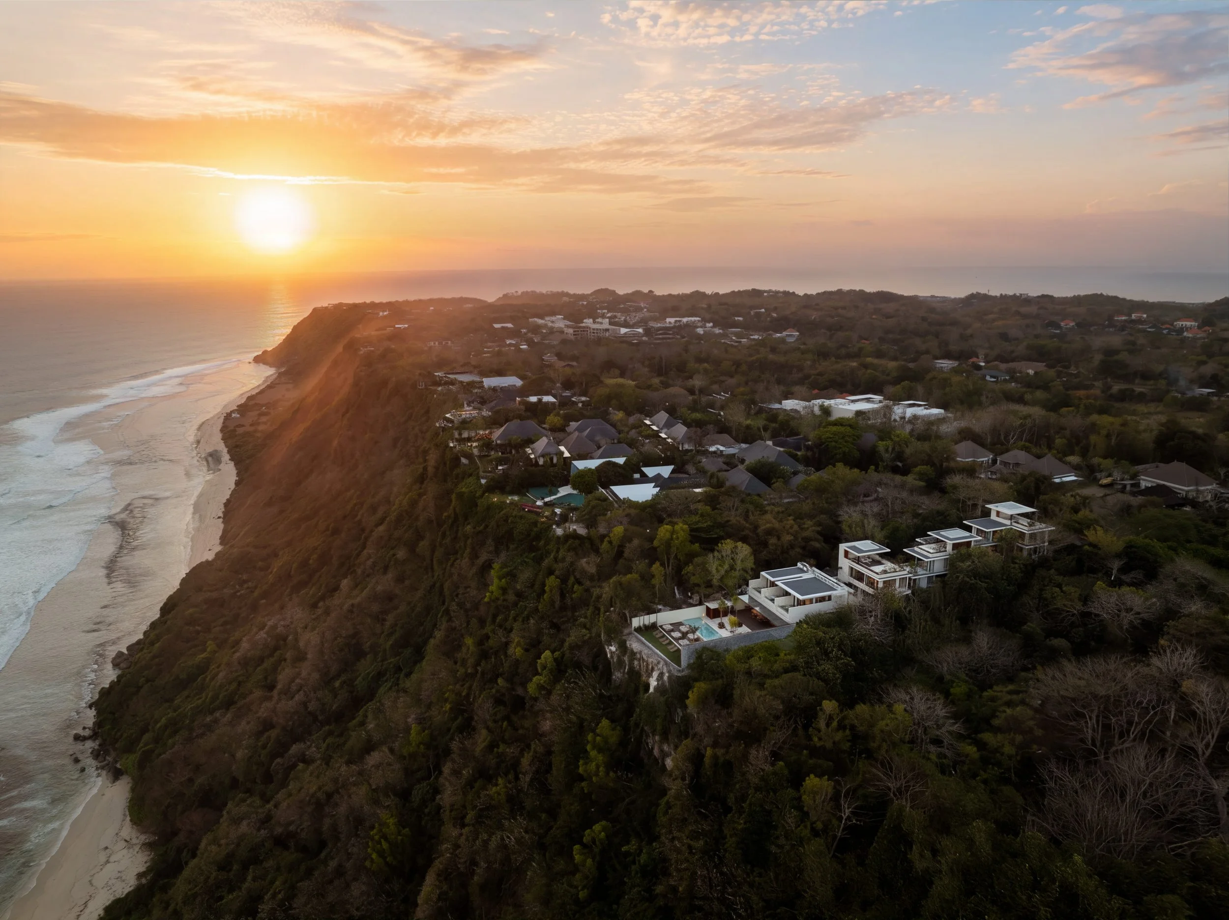 Sunset over a coastal hillside neighborhood with view of the ocean and beach.