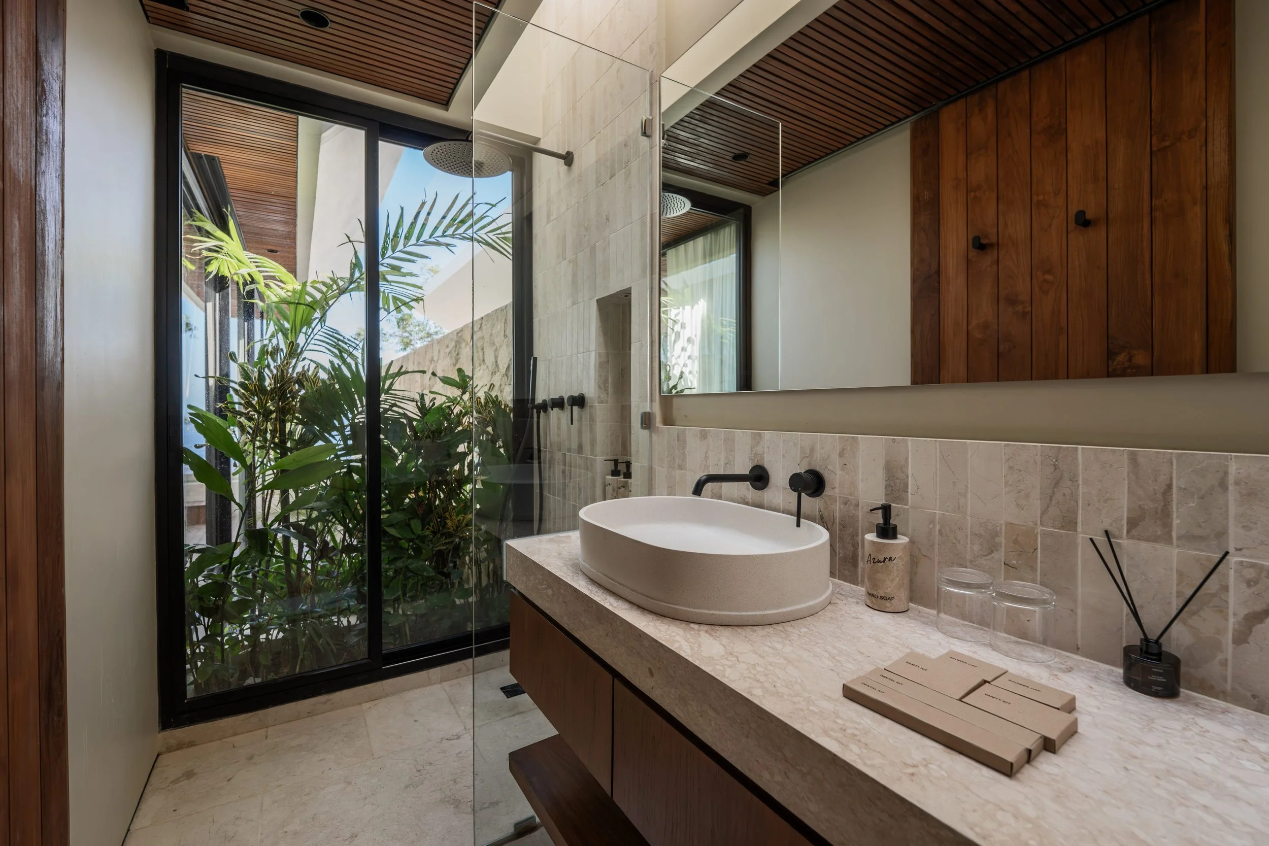 A bathroom with a large mirror above a beige stone countertop, a white vessel sink, black faucet, and a soap dispenser labeled "Azura". Two small vases with black reed diffusers and a set of folded wooden boards are on the counter. Behind the sink, t