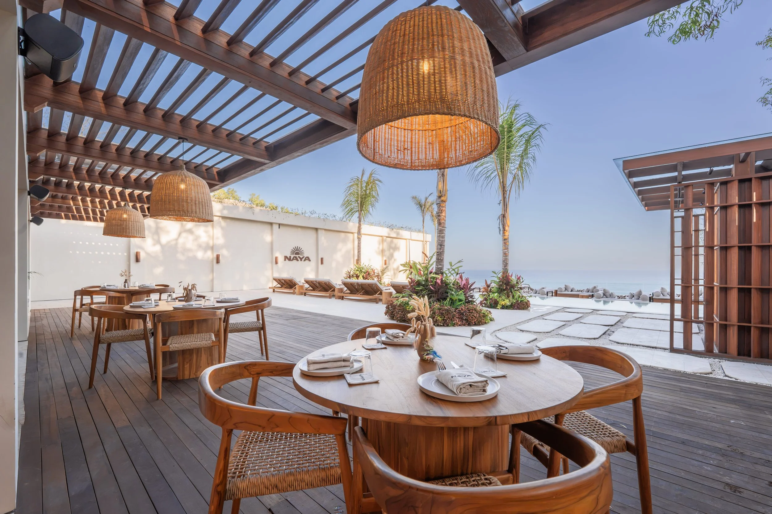 Indoor dining area with wooden tables and chairs, decorated for a meal with napkins and glasses, overlooking an outdoor area with palm trees and a view of the ocean under a clear sky.