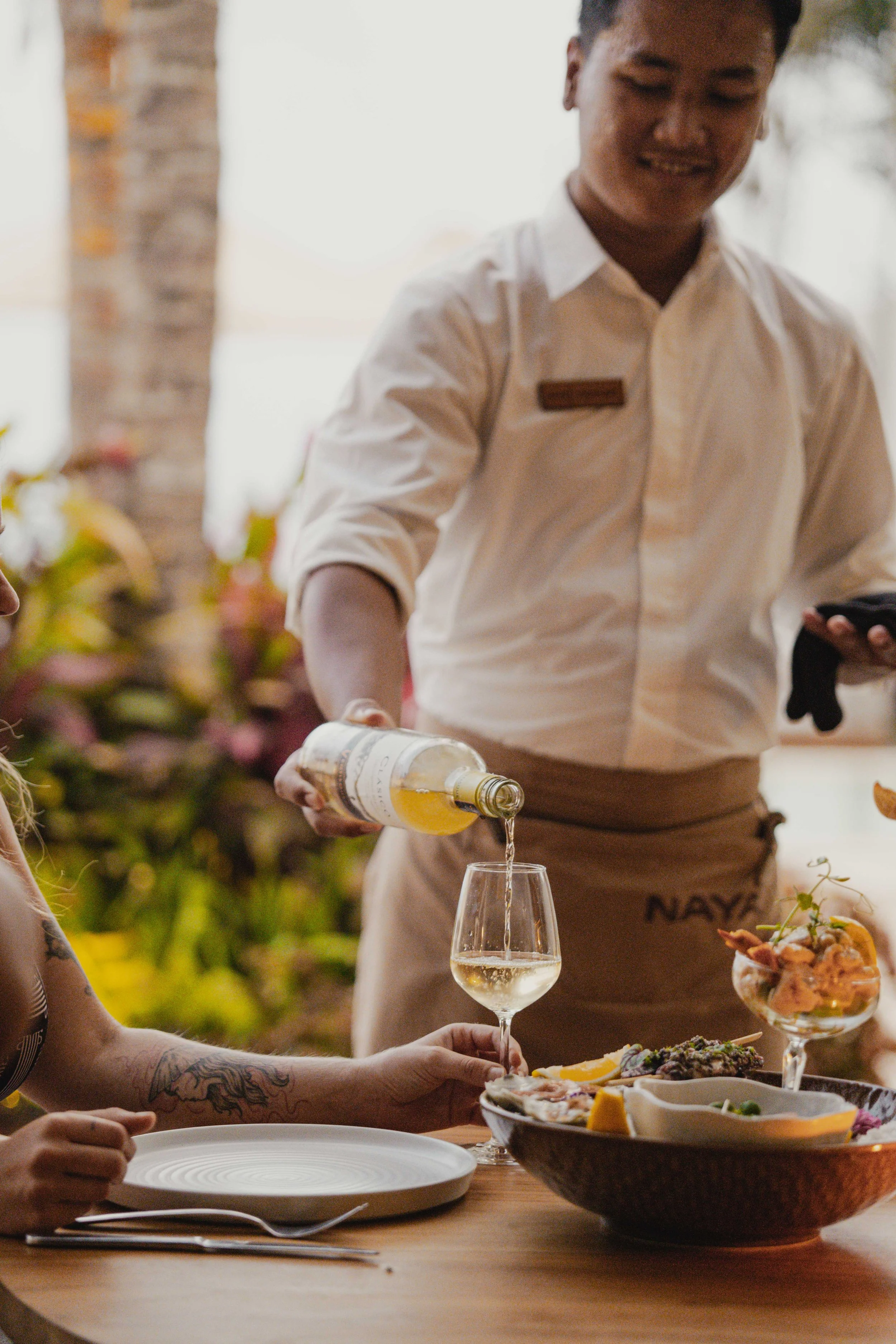 A server pouring white wine into a glass at a restaurant table, with a person reaching for food and another person holding a plate in the foreground. There are bowls of food and a plate on the table, and the background has plants and natural light.
