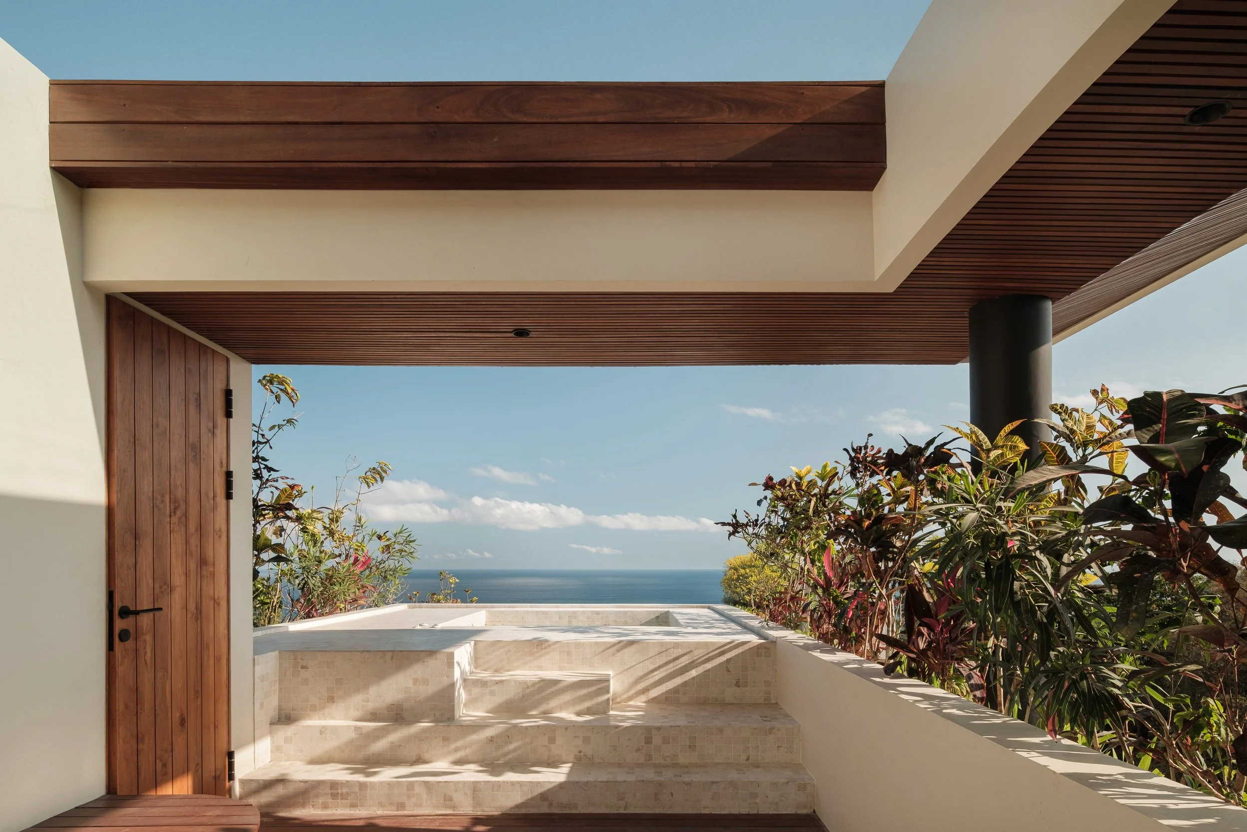Modern architectural outdoor space with a wooden door, wooden ceiling, and a view of the ocean and sky in the background.