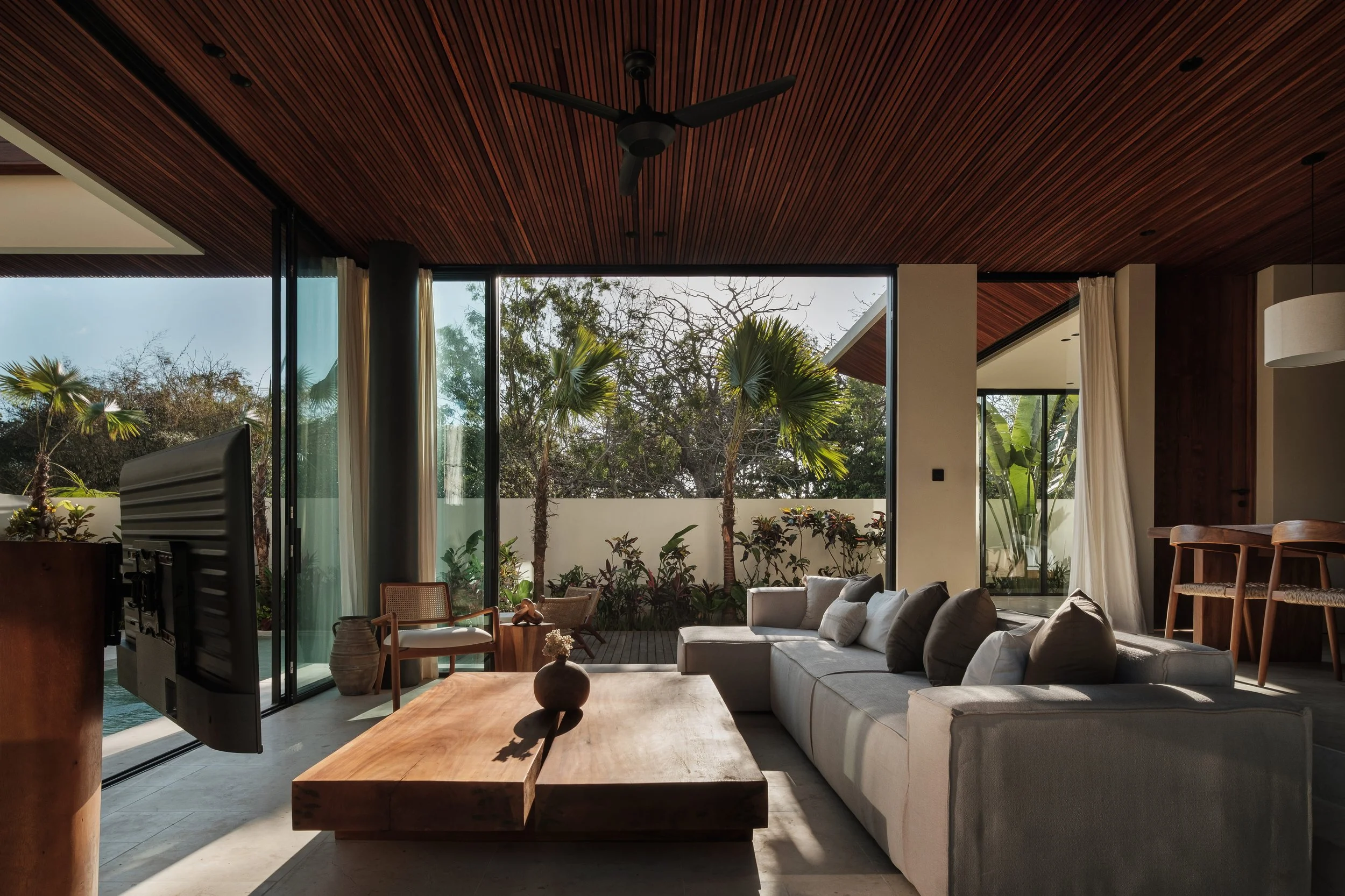 Modern living room with large glass windows showing outdoor trees, beige sectional sofa, wooden coffee table, and ceiling fan with wooden slats.