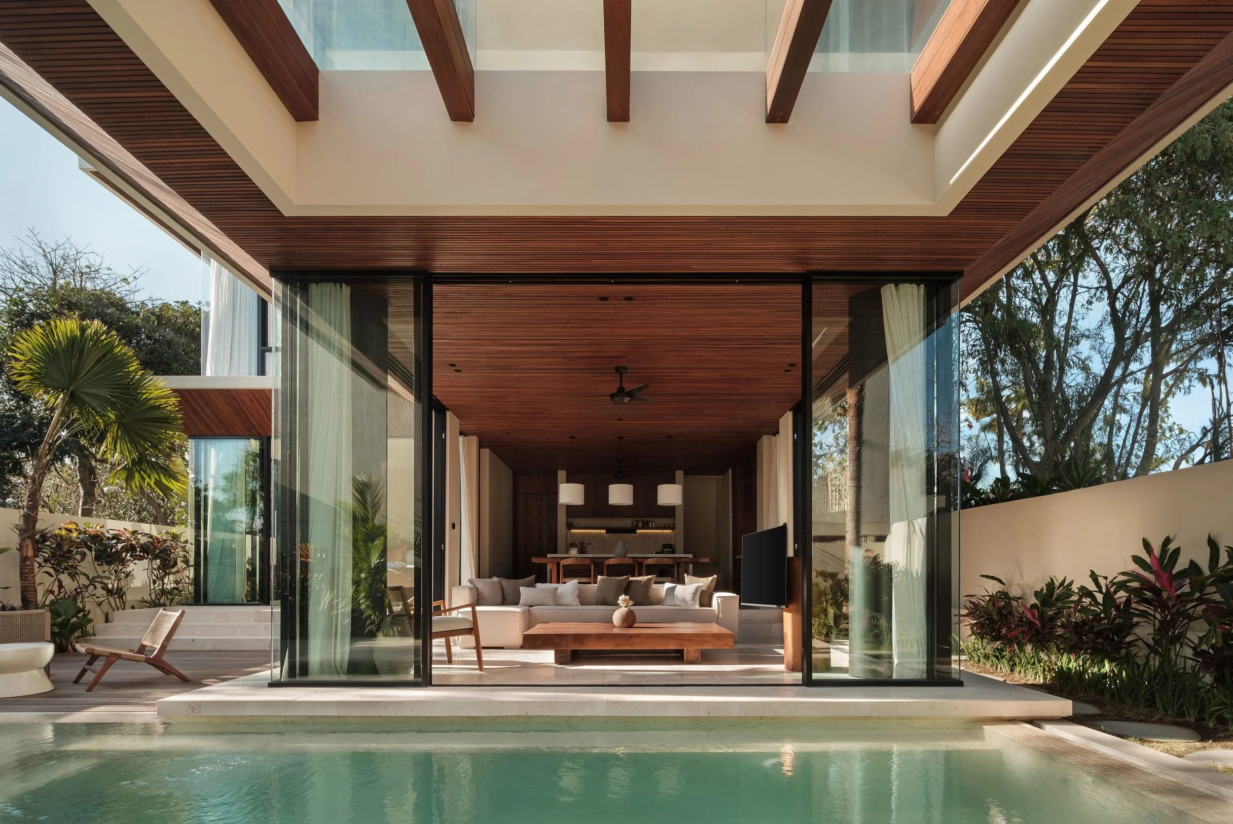 Modern open-concept house with large glass sliding doors opening to a pool and tropical landscaping.
