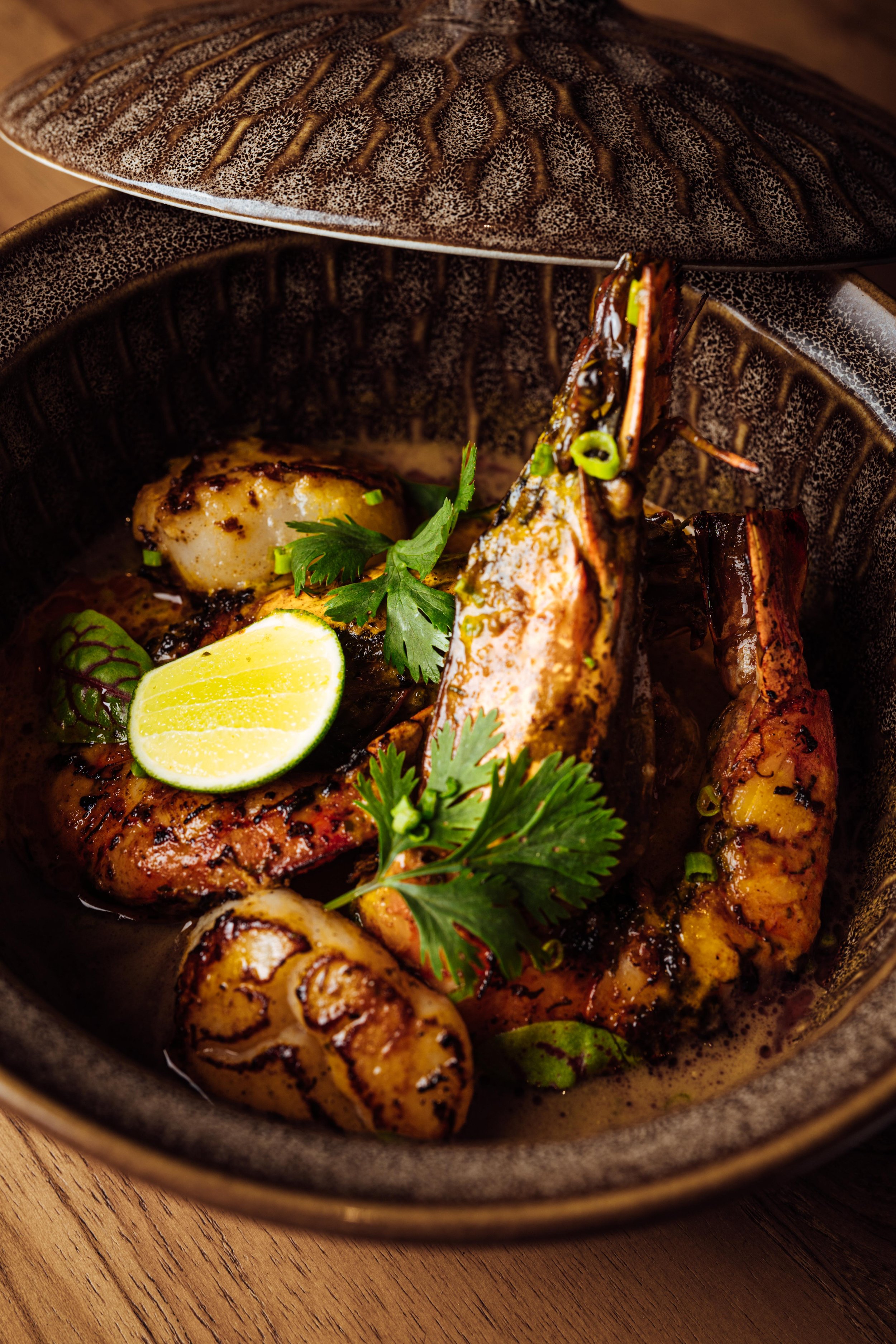 Close-up of grilled seafood, including lobster tail and scallops, garnished with lime and fresh herbs in a dark bowl on a wooden surface.